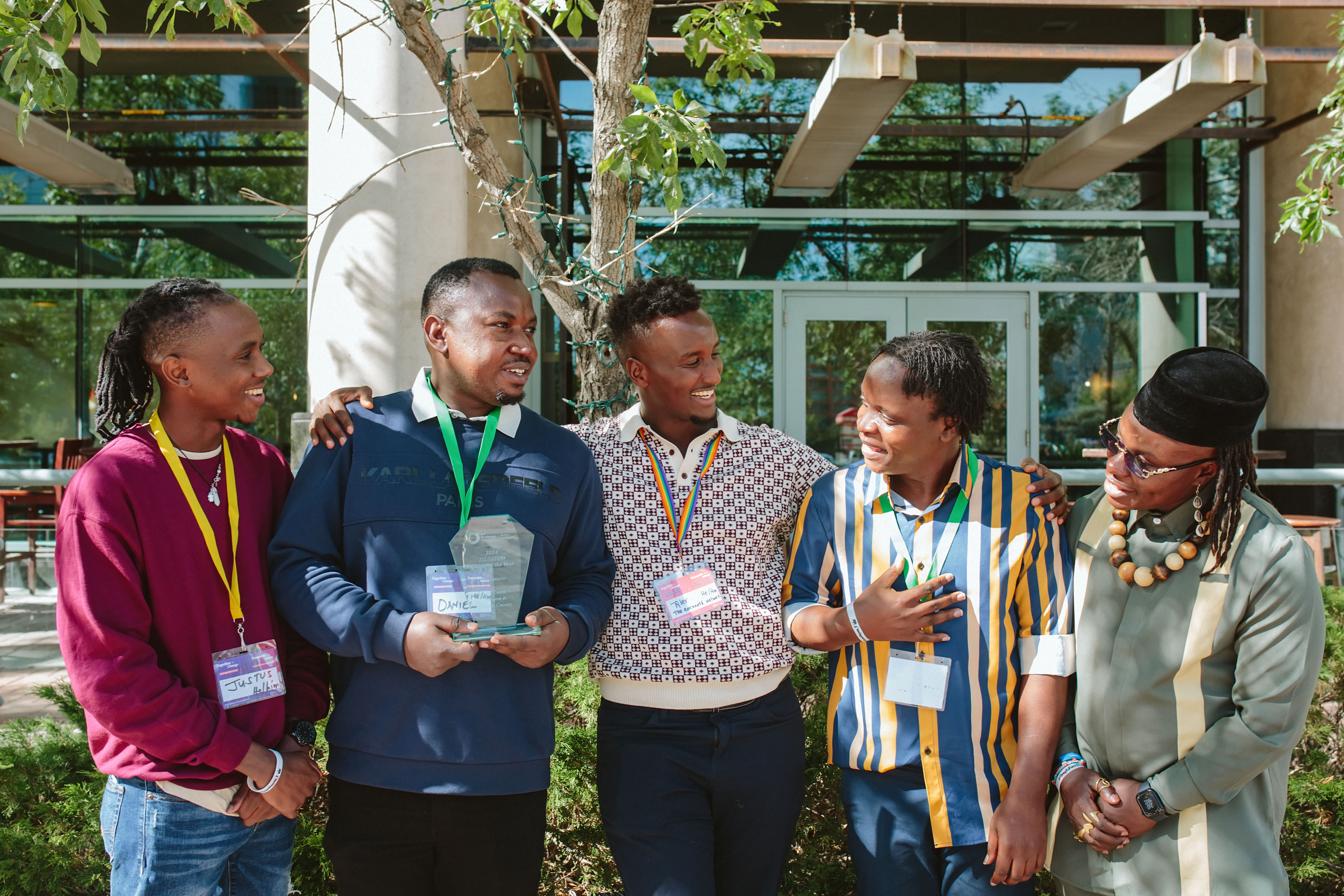A group of five people smiles together outdoors, holding items and wearing name tags, in a vibrant setting.