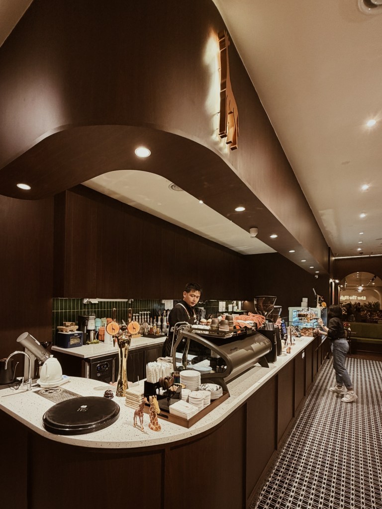 Interior of a coffee shop with a dark wooden counter, a barista preparing drinks, and a customer browsing pastries. The decor includes unique accessories and stylish lighting.