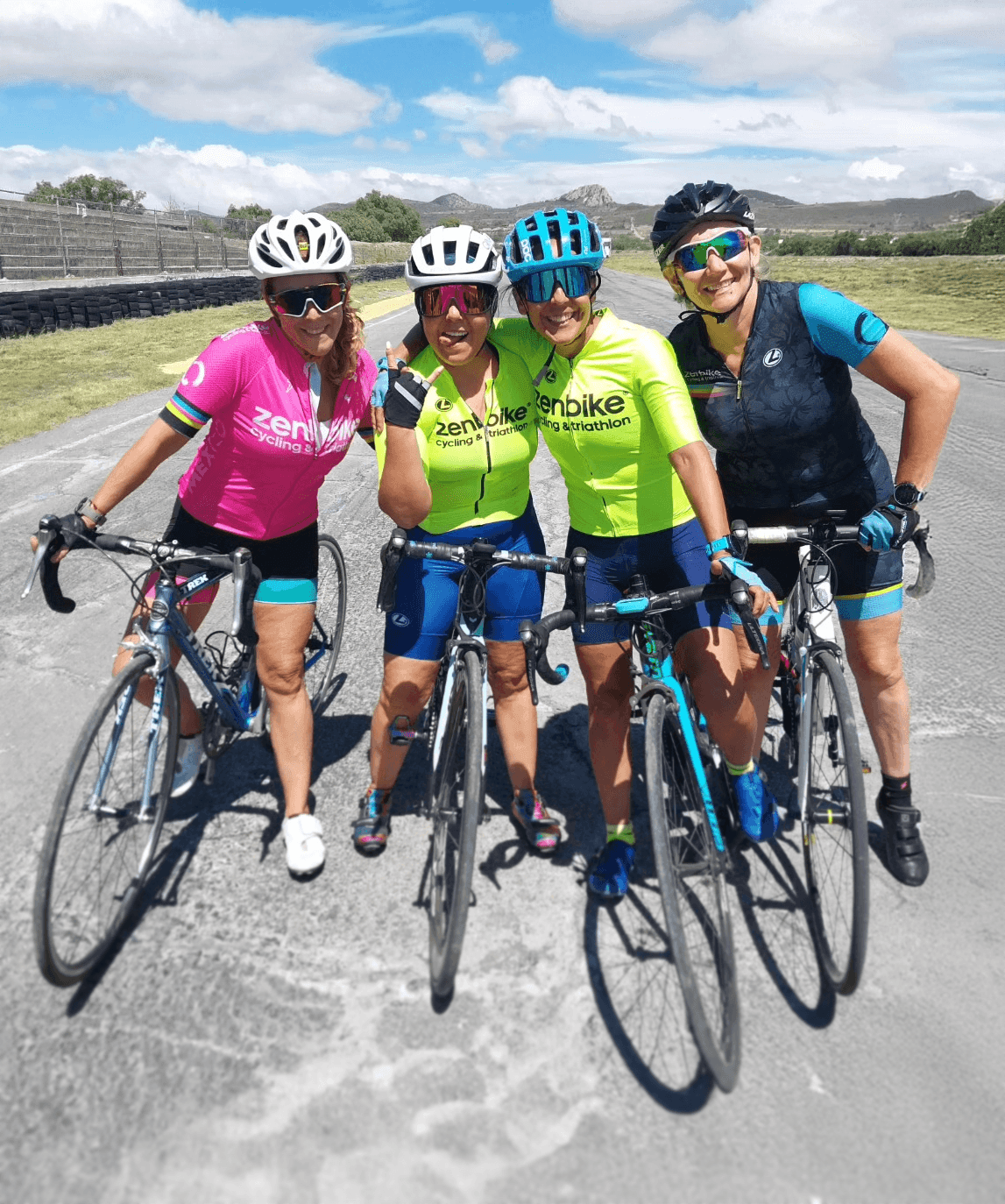 Ciclistas zenbike posan sonrientes en pista al aire libre, luciendo uniformes de diferentes colores bajo cielo despejado.