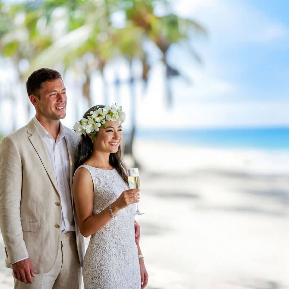 Smiling couple in wedding attire with champagne on a tropical beach at Uprising Beach Resort.