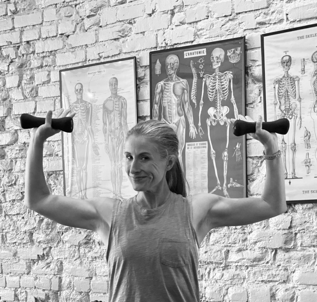 A person exercises with dumbbells in a black and white image, posed in front of anatomical posters on a brick wall, showcasing fitness and health themes.