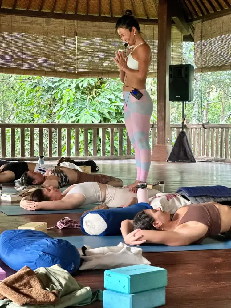 Vivienne Zeng leading a yoga teacher training session in a Bali shala, students resting in Savasana with bolsters.