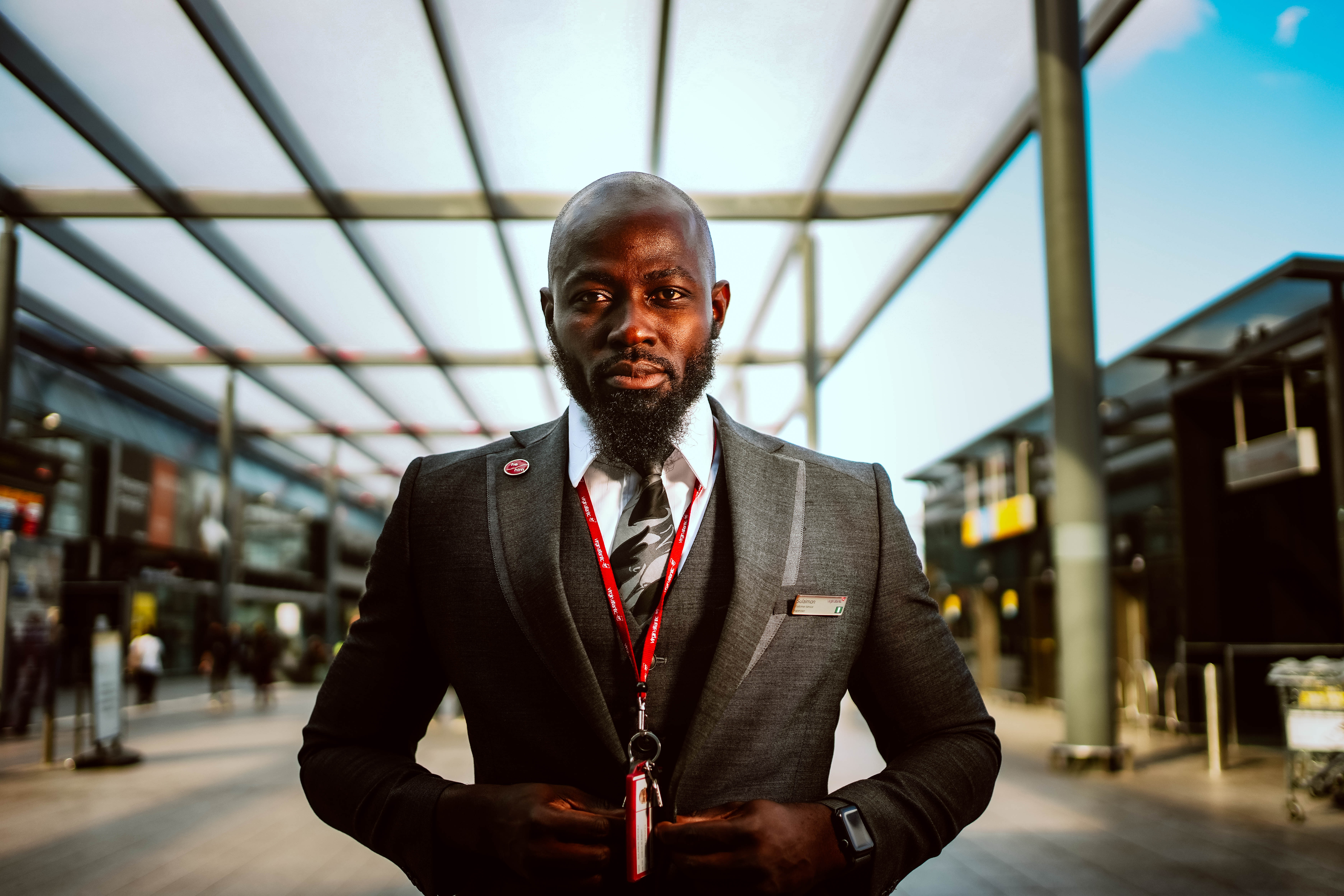 A powerful, low-angle portrait of a Black man with a beard, dressed in a professional suit and tie, looking confidently into the camera while standing under a modern outdoor canopy.