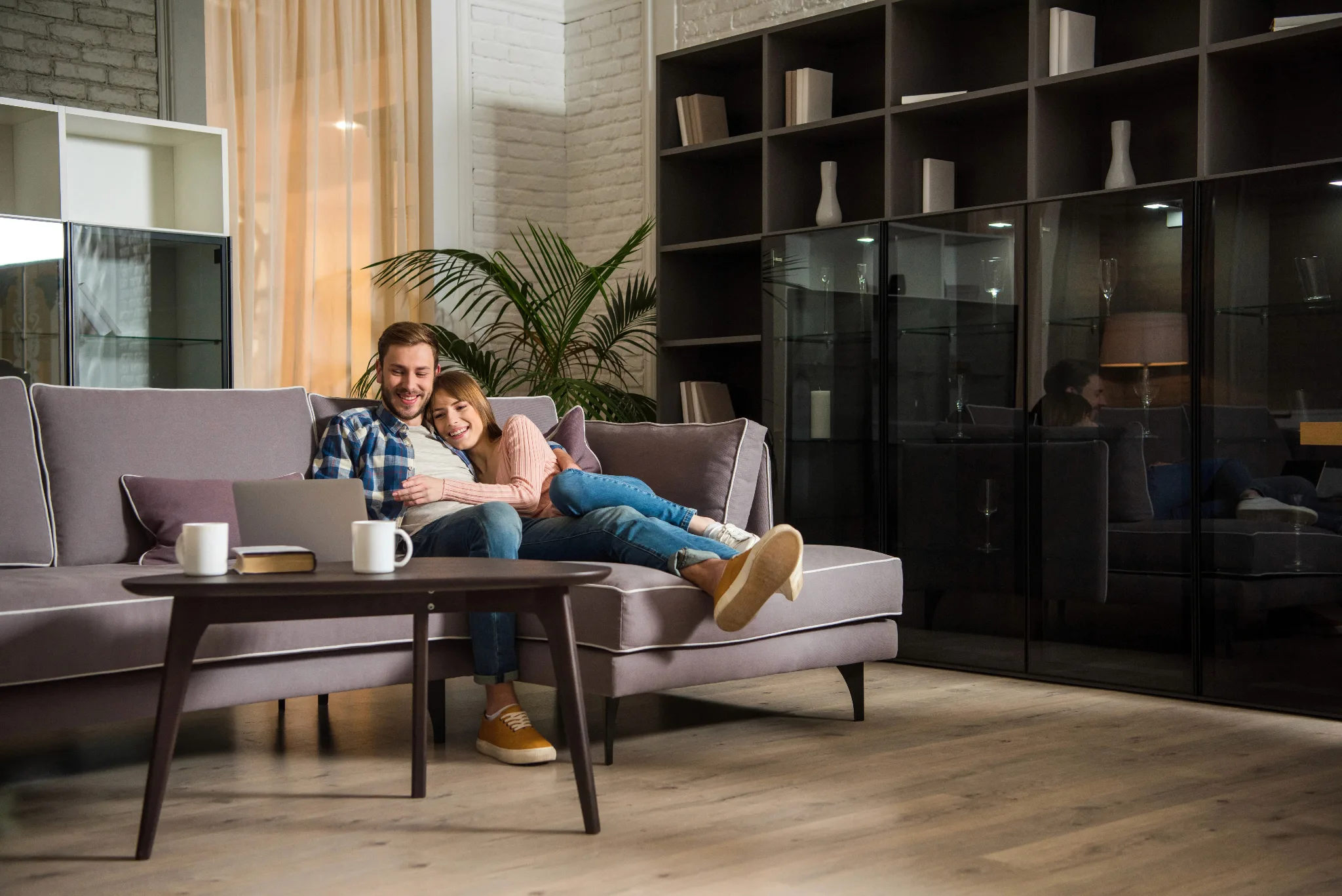 Couple lying on a couch, female hugging the male, both looking at a laptop placed on the center table, a big wooden cabinet at the back