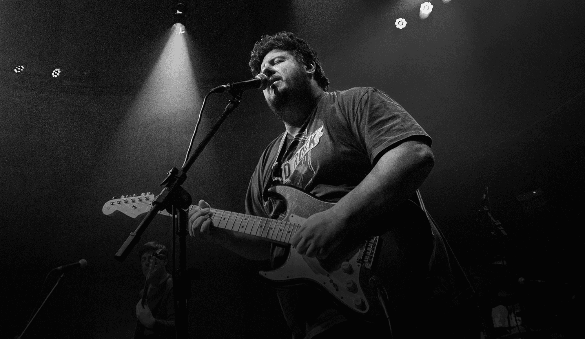 Guitarrista de blues internacional se apresentando ao vivo no palco, cantando ao microfone e tocando guitarra durante show.
