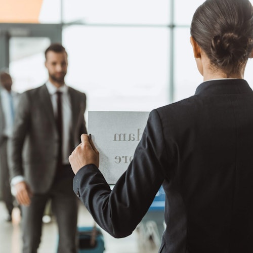Una persona in abito che tiene un cartello all'altezza della vita di fronte a un'altra persona in abito grigio che cammina verso di loro in un aeroporto.