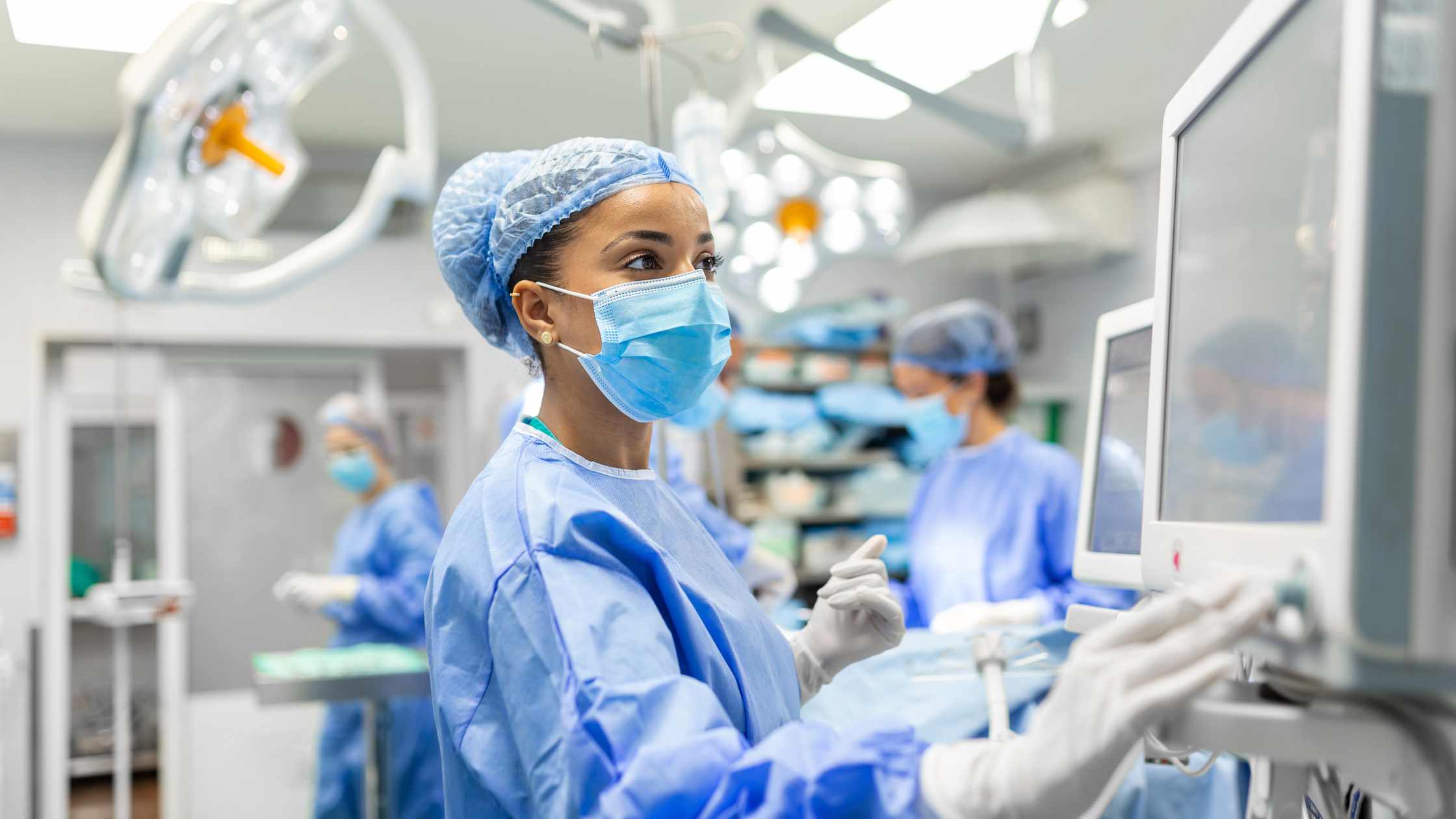 Masked surgical nurse in blue scrubs looking at a monitor in an operating theatre, with colleagues working behind