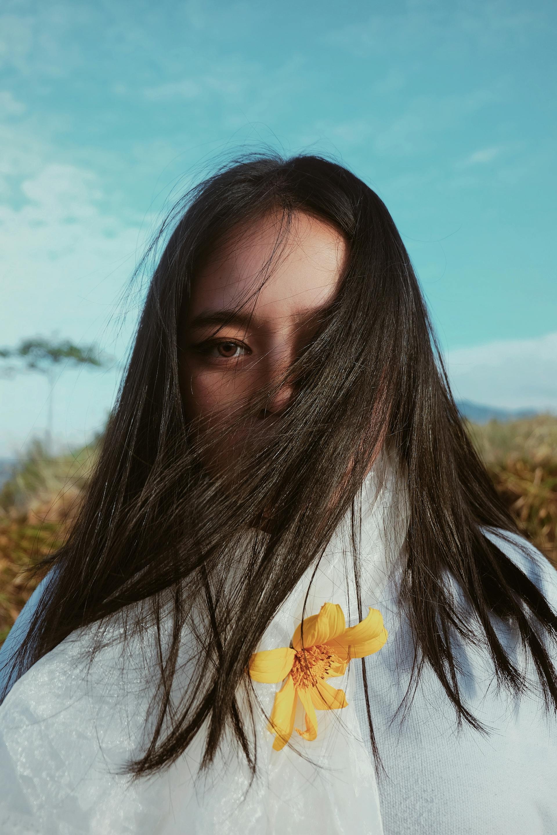 A close-up shot of a woman with long dark hair blowing across her face, revealing one eye. She wears a white shirt with a single yellow flower tucked in, set against a blue sky.