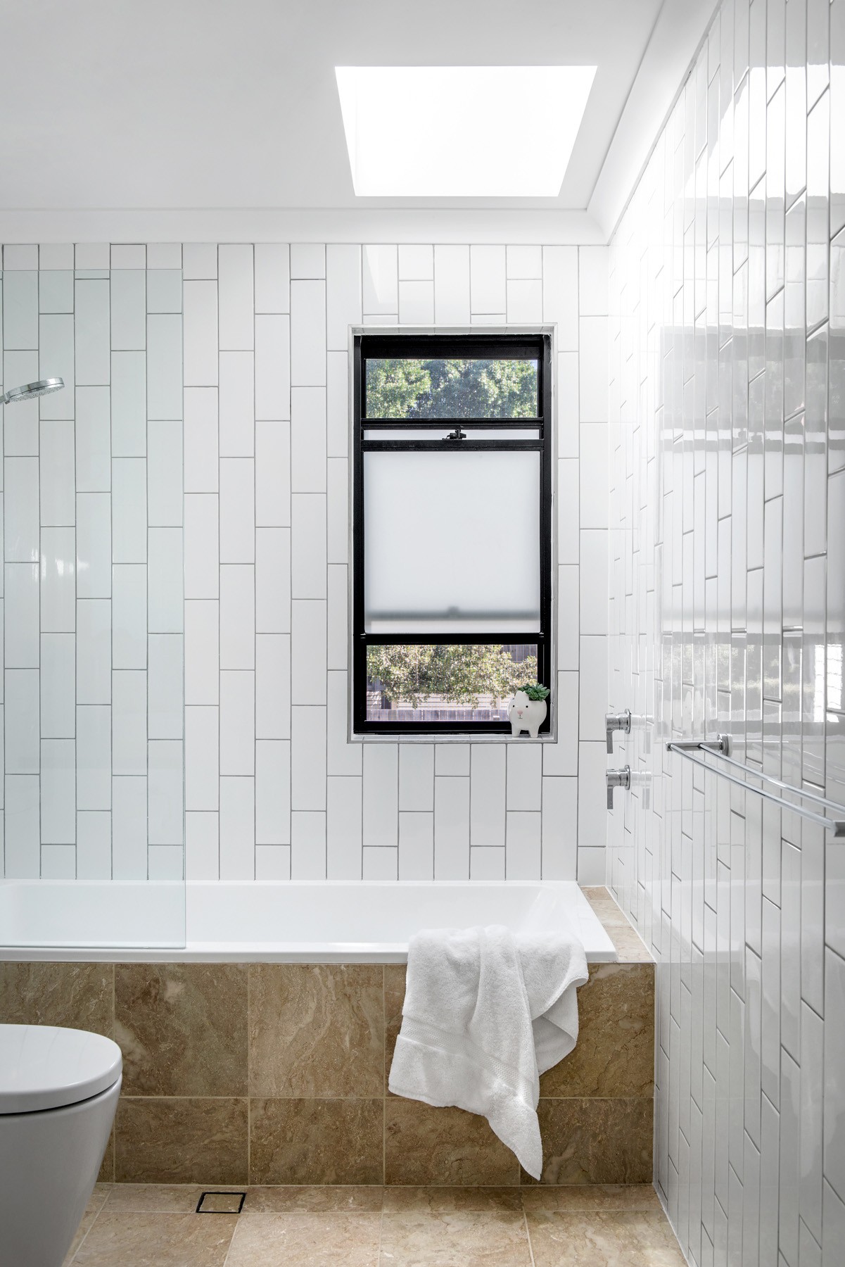 Bathroom at Oxford Cottage with white tiled walls, a skylight over the bath, and warm stone finishes grounding the space.