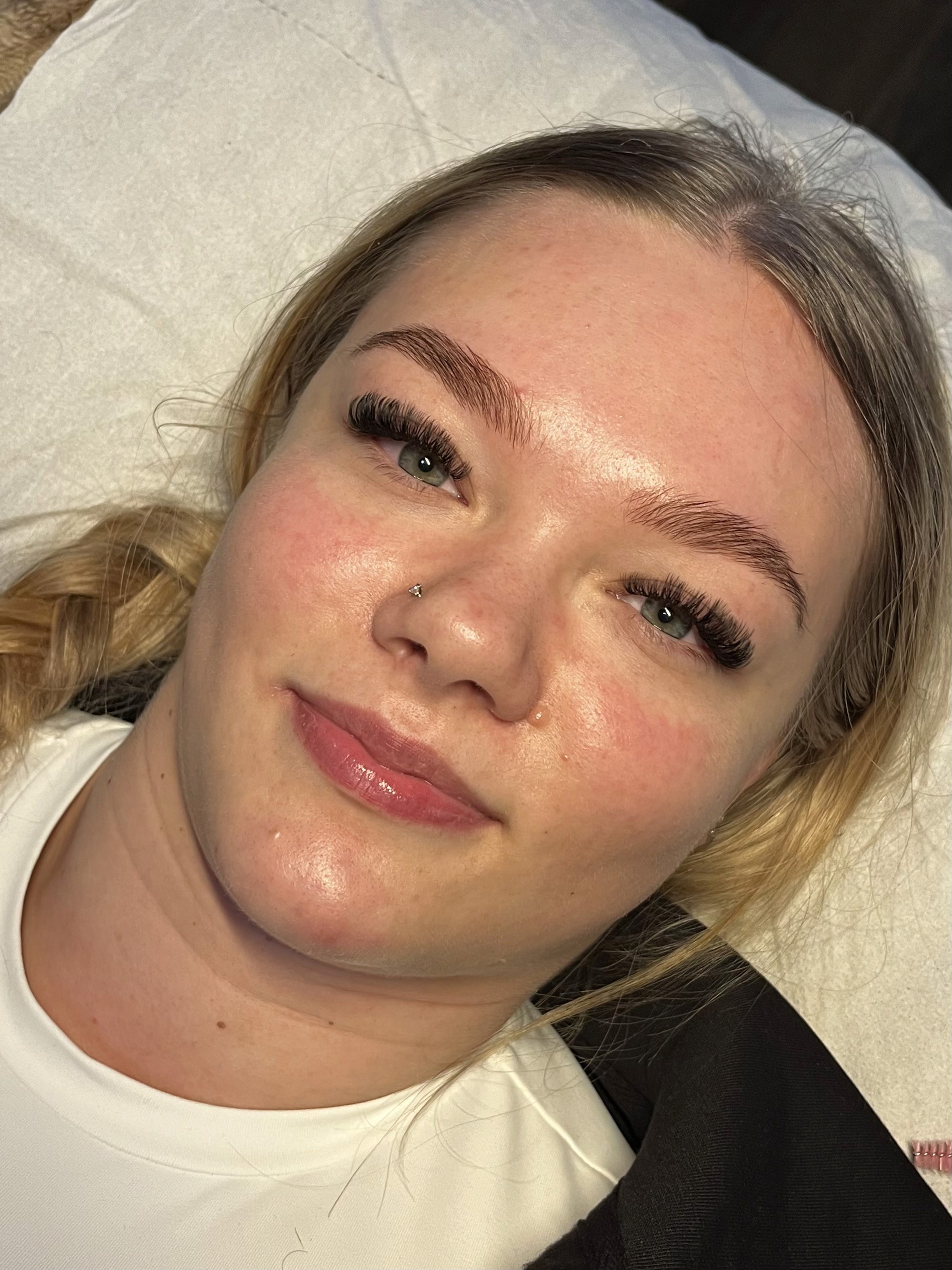 Woman with braided blonde hair and false eyelashes, smiling slightly while lying on a pillow. Her skin appears smooth and well-lit.