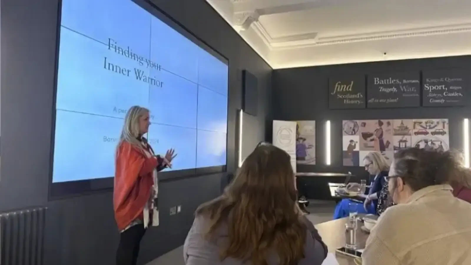 Woman presents "Finding Your Inner Warrior" on a large screen in a conference room. Attentive audience of women listens, fostering an empowering atmosphere.
