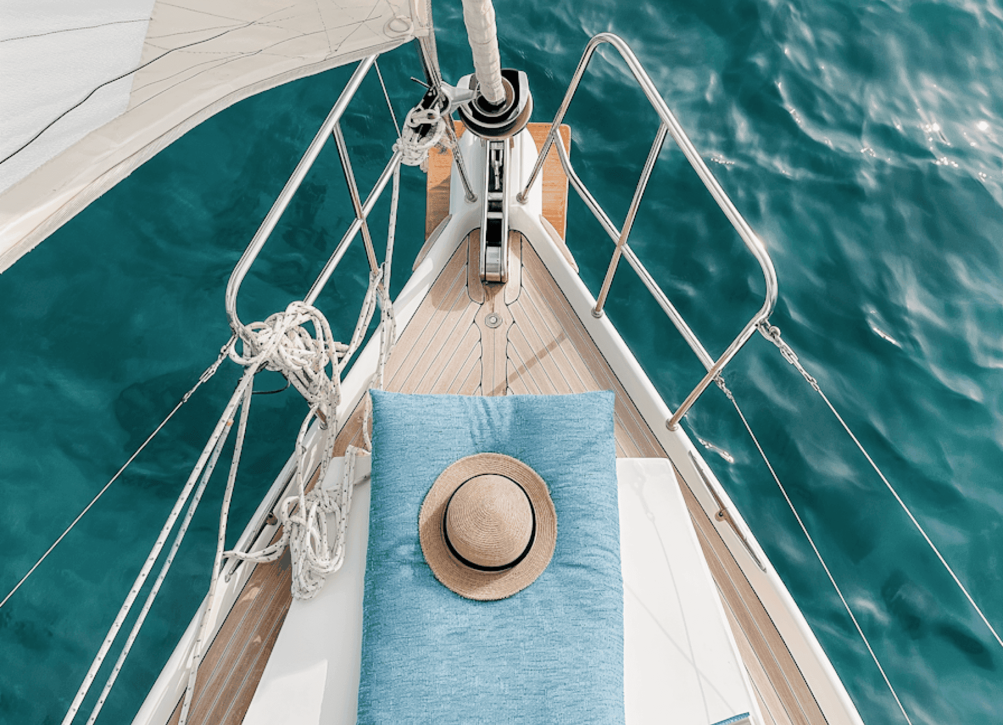 Relaxing spot on a sailboat deck with a straw hat on a cushion above calm turquoise water.
