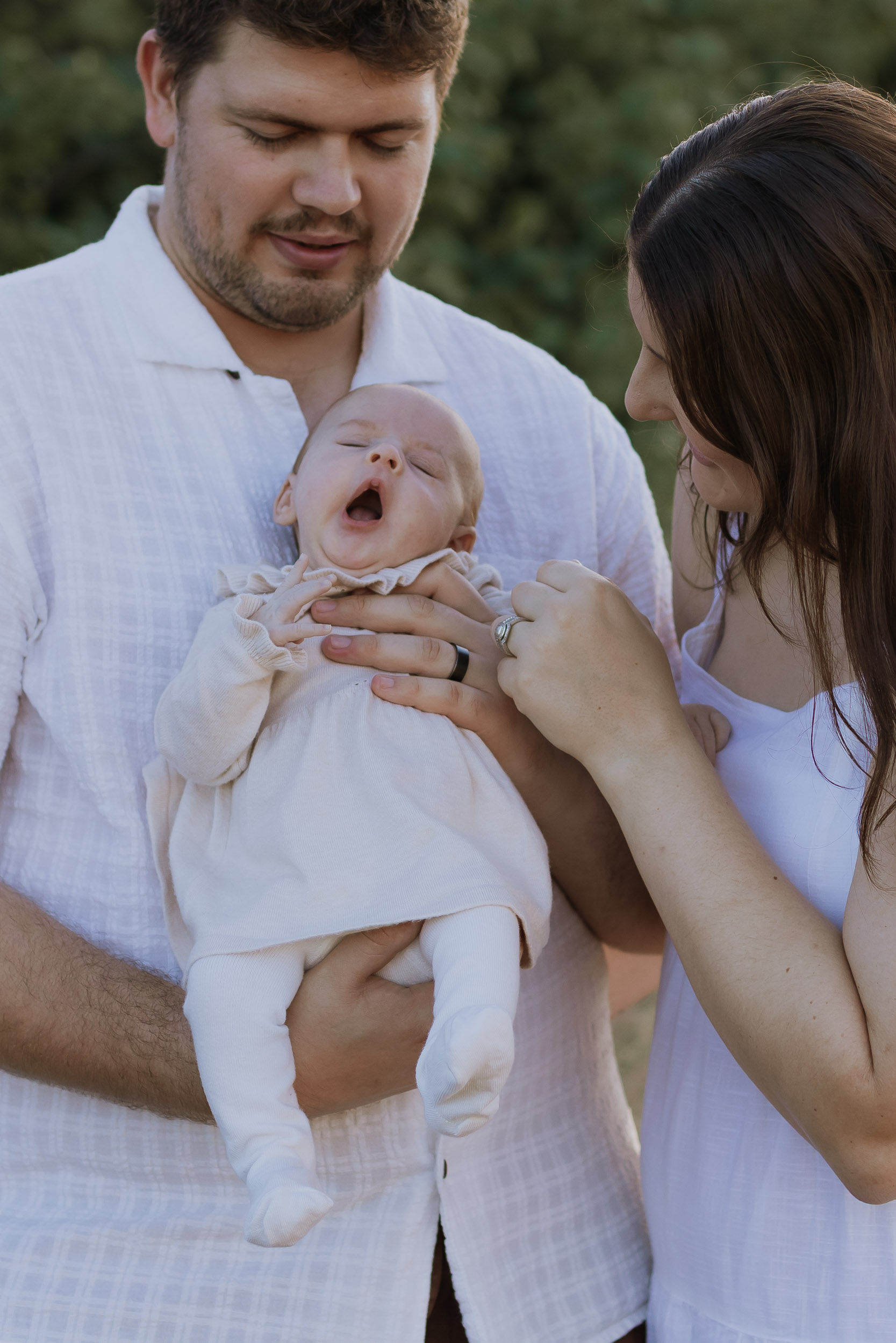 Parents holding yawning newborn baby in grassy outdoor photoshoot