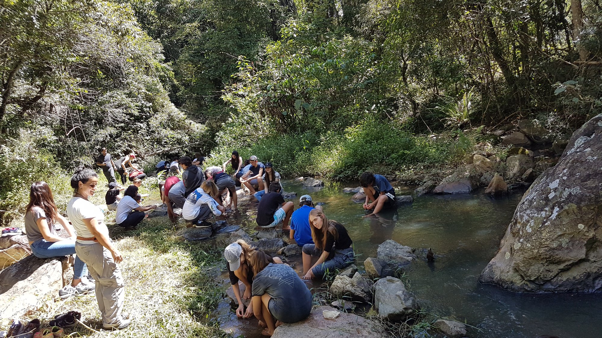 Grupo grande de alunos sentado nas margens e pedras de um rio cercado de mata