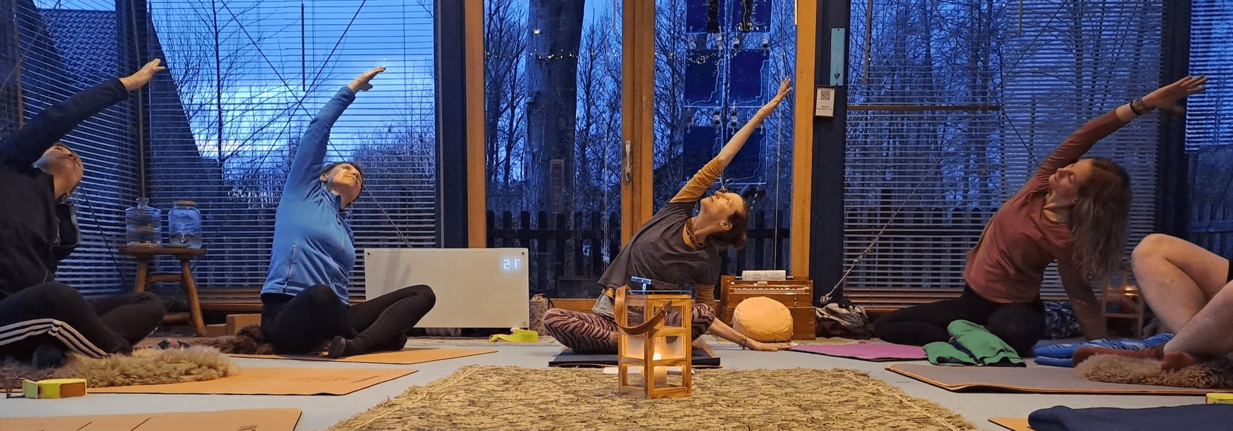 4 Frauen sind auf Yogamatten in einem Raum mit großen Fenstern in einer Seitbeuge im gekreuzten Sitz (Aufwärmübung)