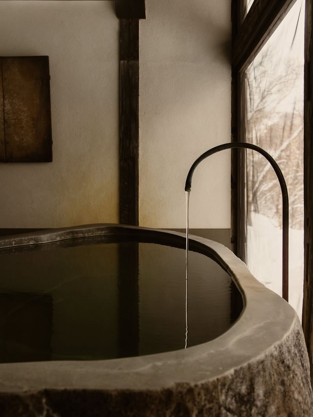 Minimal Japanese bath with a dark soaking tub, curved faucet, and snowy window view.