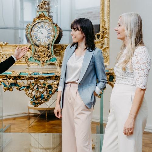 Zwei Frauen stehen in der Nähe einer kunstvollen antiken Uhr, während eine dritte Person in ihre Richtung deutet. Die Szene scheint sich drinnen abzuspielen.