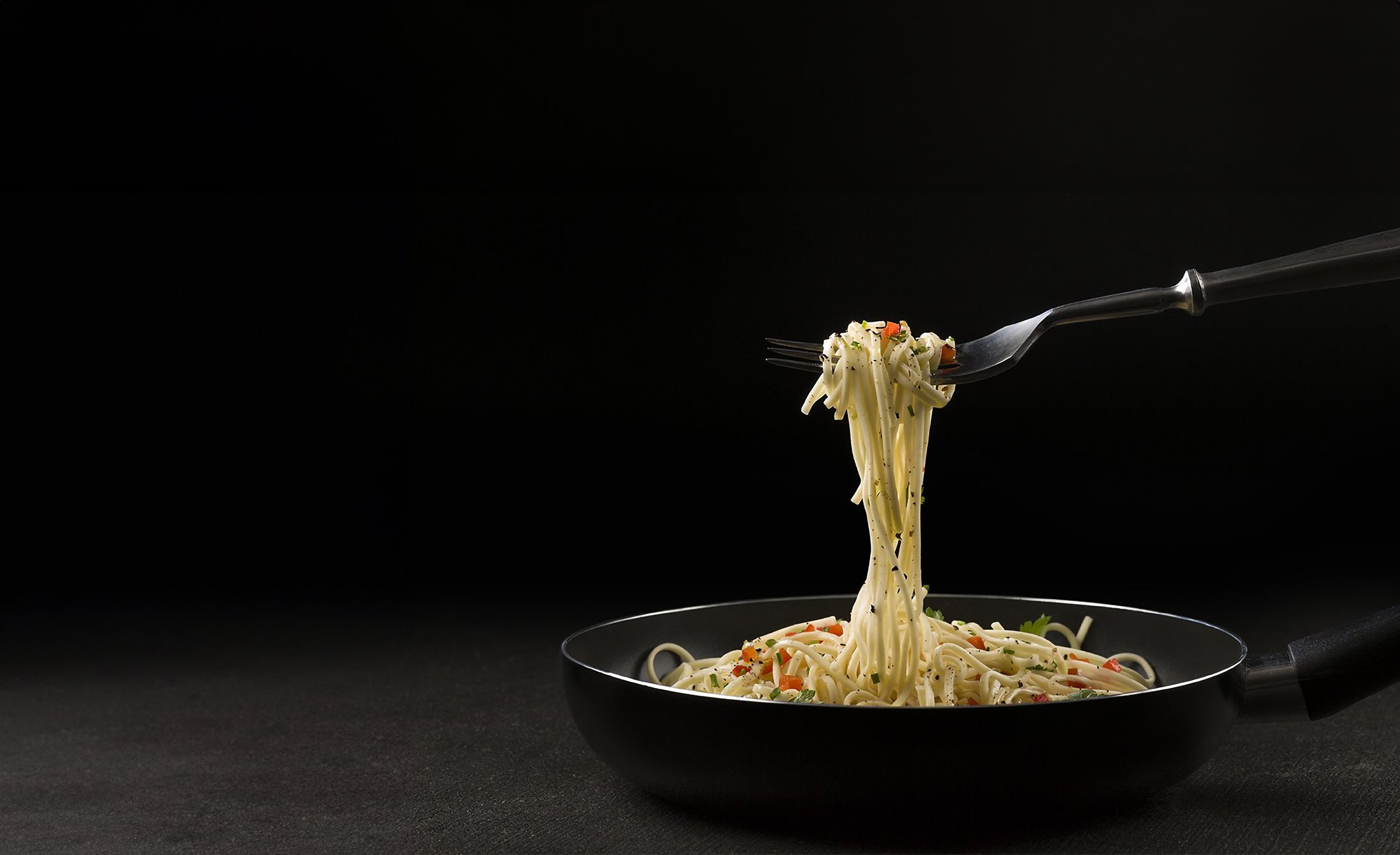 A fork lifts spaghetti from a dark skillet against a black background. The noodles are garnished with herbs and small colorful vegetables, creating a gourmet feel.