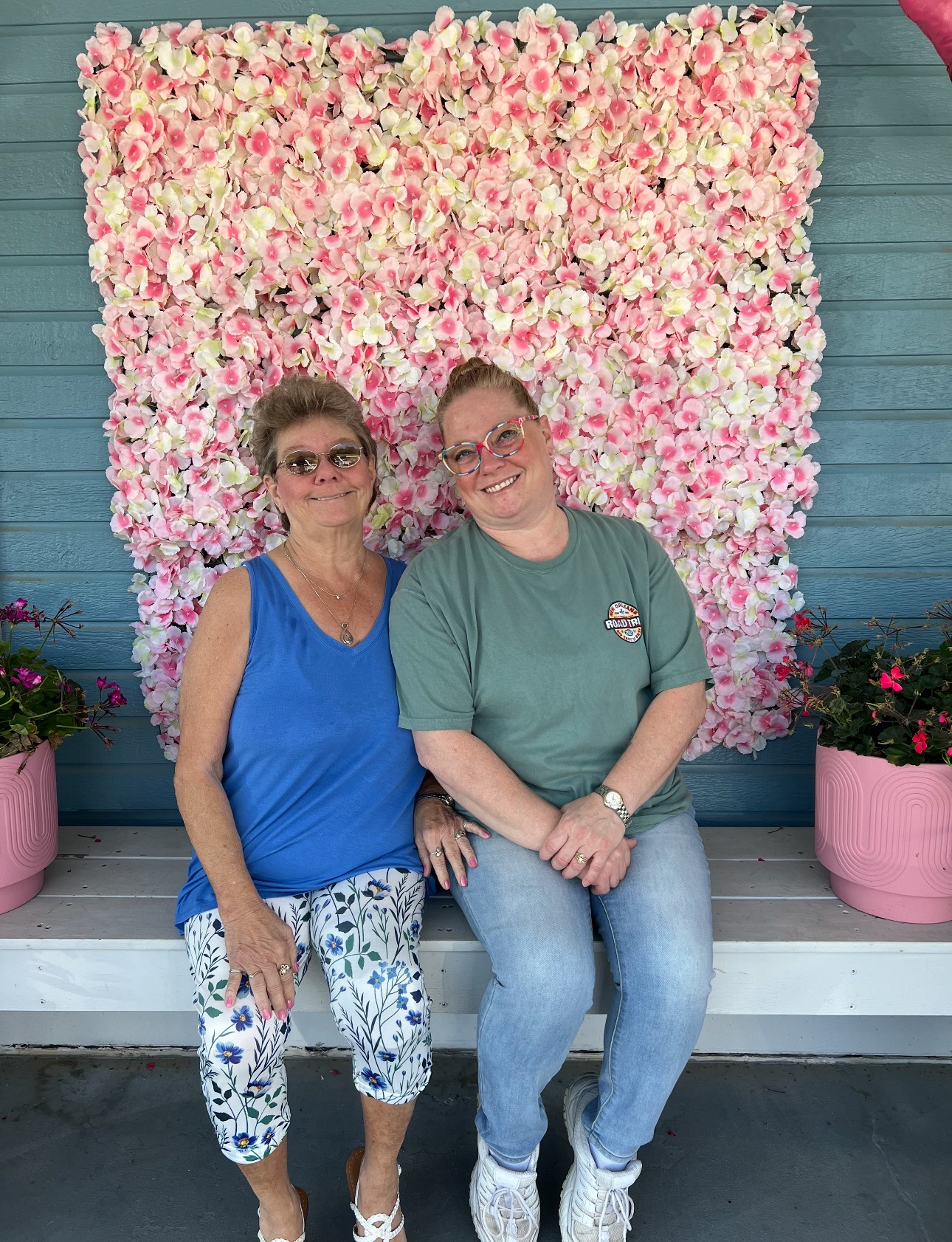 Two individuals sit on a white wooden bench in front of a vibrant pink flower wall, flanked by pink flower pots and against a blue paneled backdrop, creating a cheerful and colorful scene.