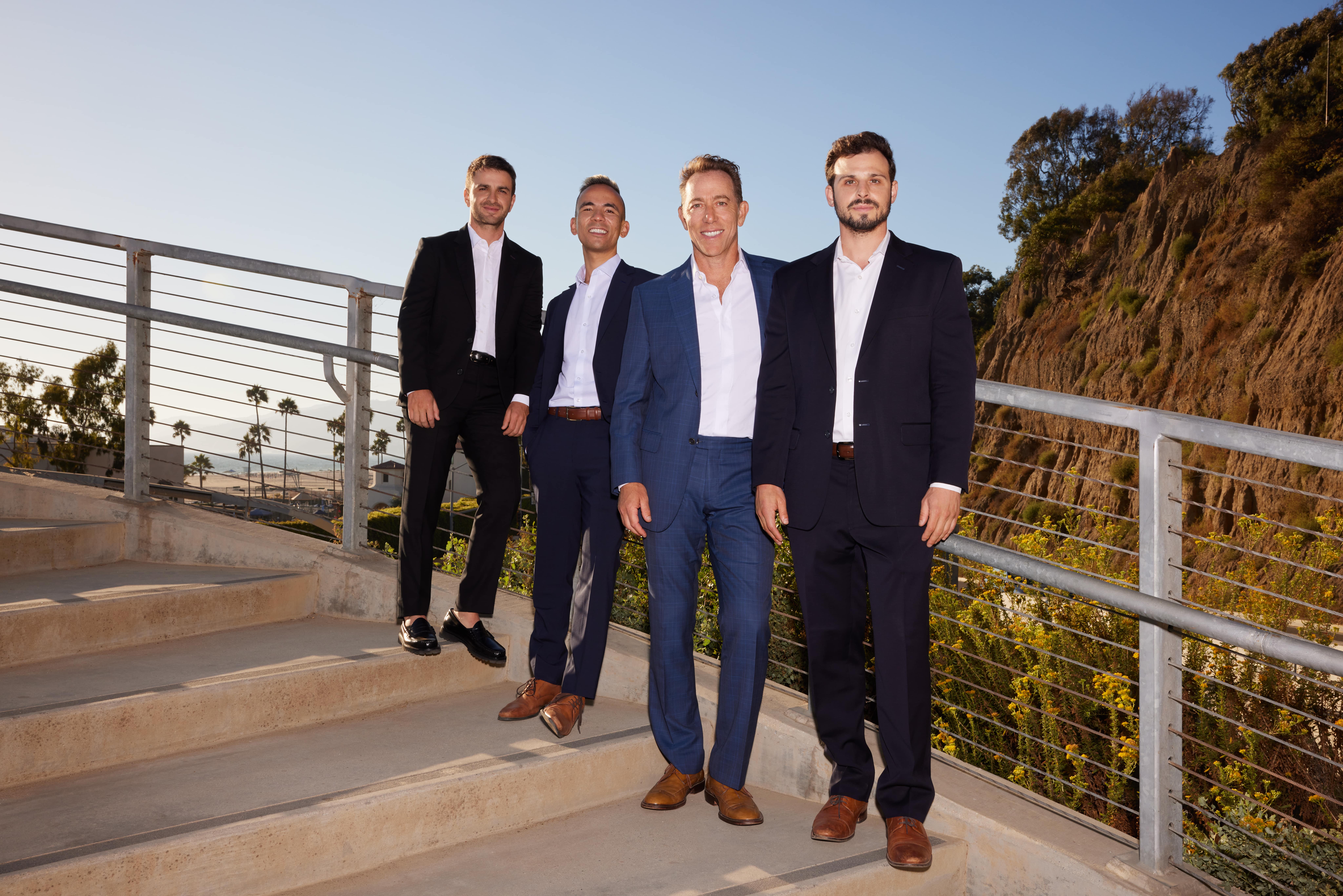 Kenny Stevens Team in business suits stand together outdoors on a pathway, with greenery and a clear sky in the background.