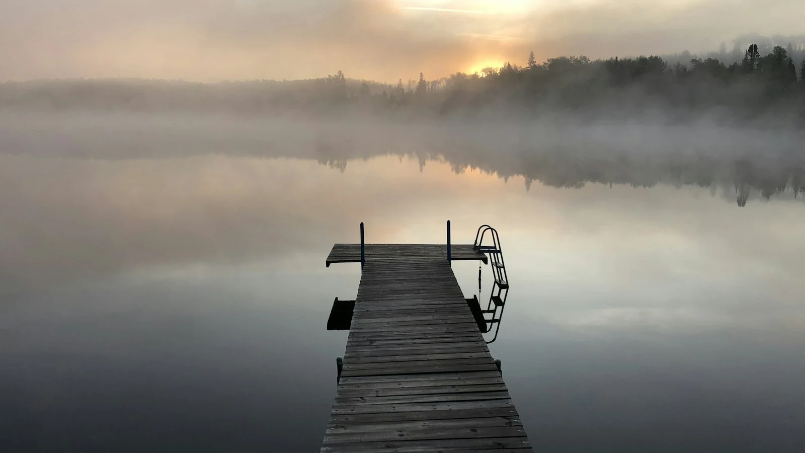 Ein hölzerner Steg ragt in einen stillen See, über dem Morgennebel liegt