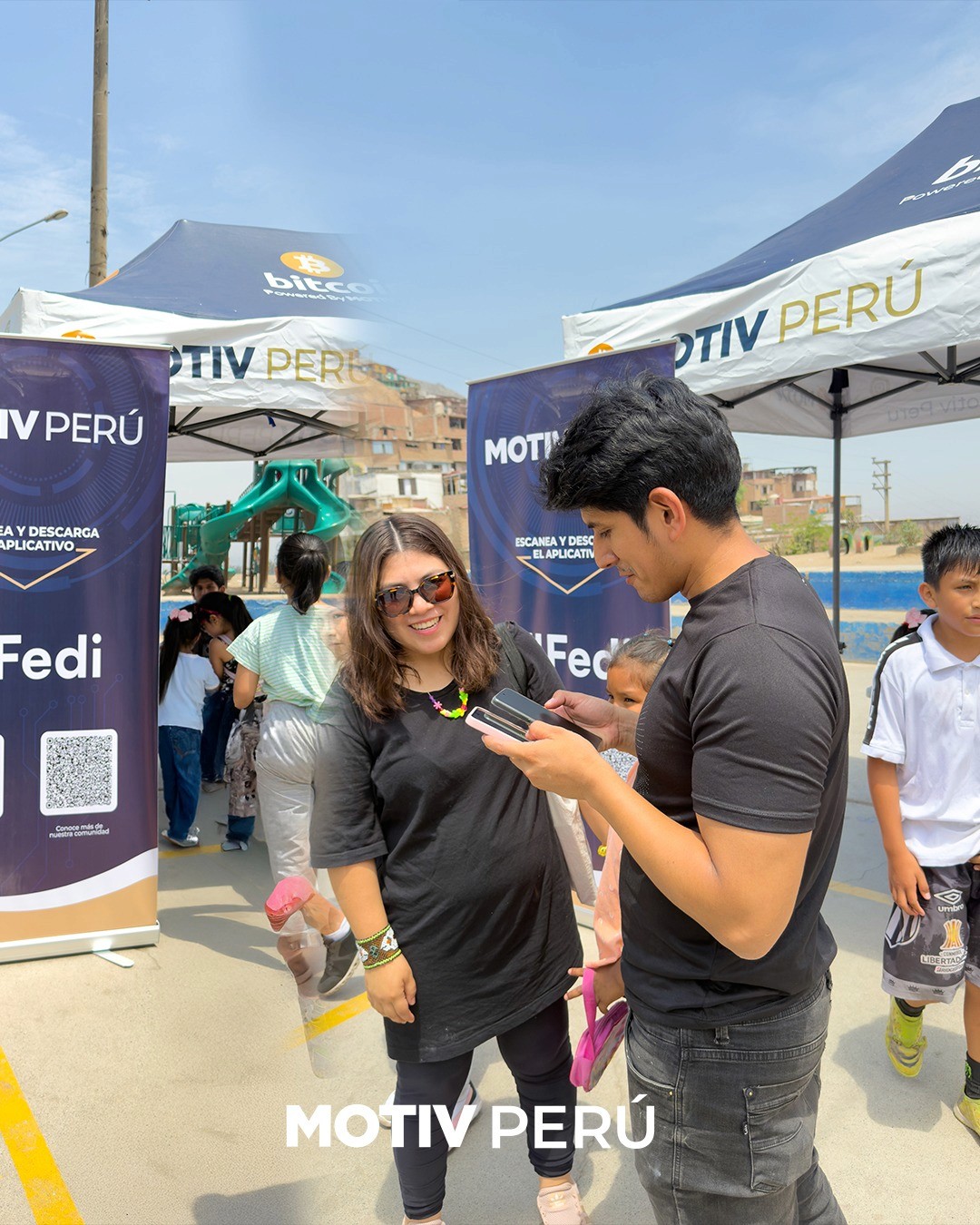 A community member gets help setting up the Fedi app at a Motiv Perú outdoor event, surrounded by Motiv and Fedi branded banners and Bitcoin gazebos. Children and families fill the background of the busy public gathering.