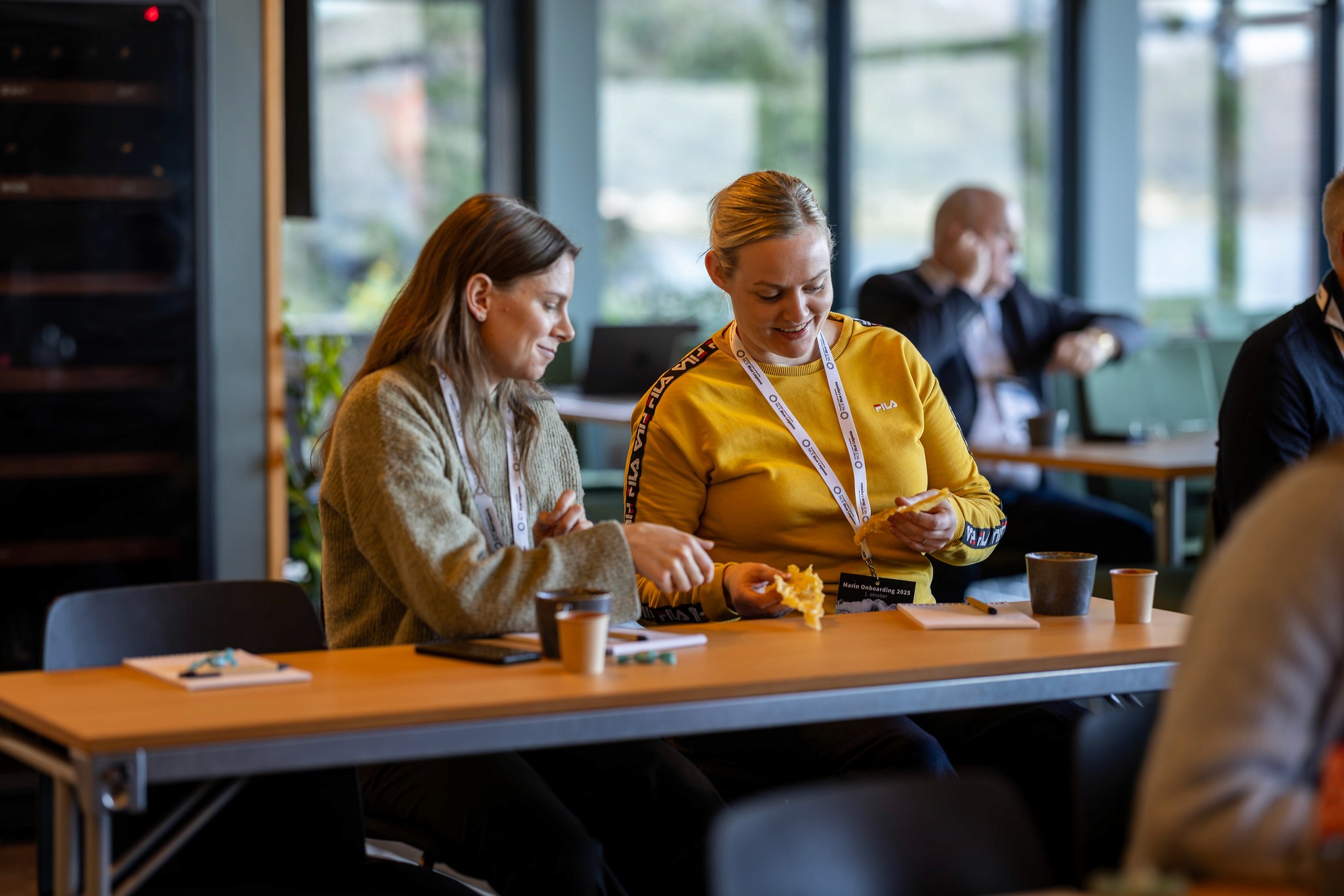 Two people sit at a wooden table in a bright room with large windows, engaged in conversation.