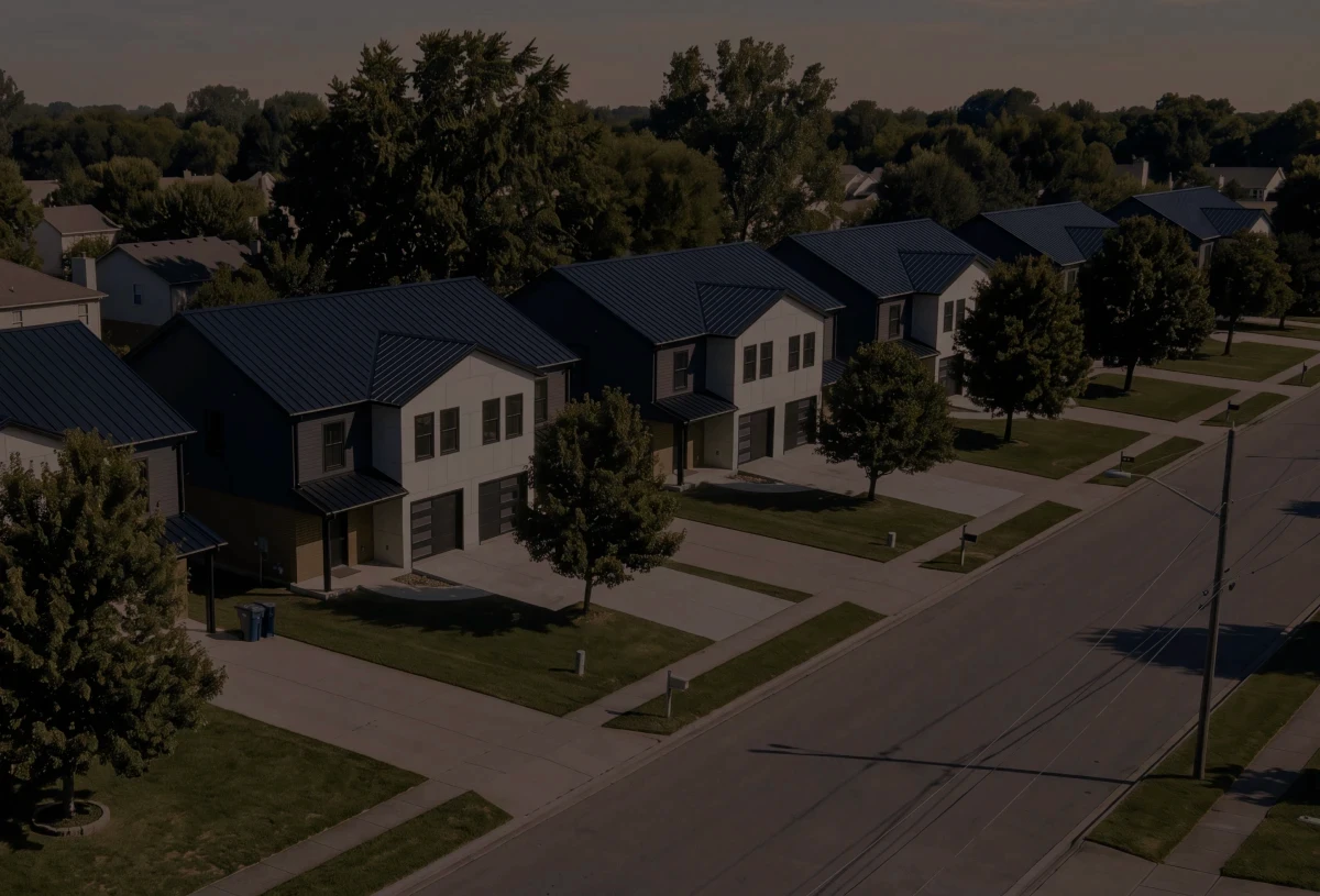 Aerial view down a street of newly built two-story homes with dark standing-seam metal roofs, attached garages and mature trees