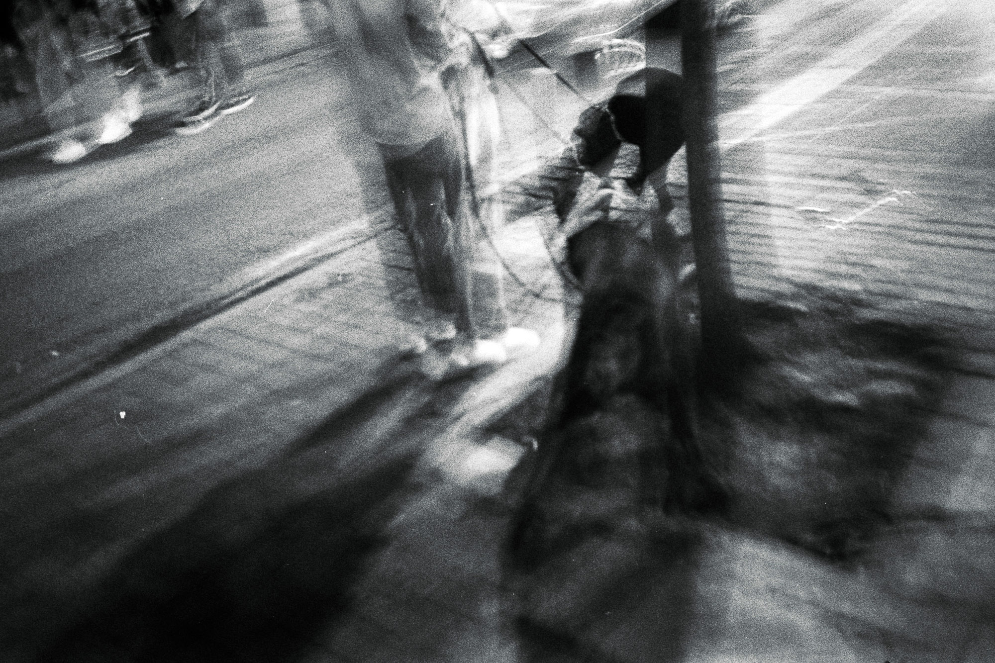 A black and white image captures a blurred street scene with two individuals and a dog on a leash, conveying a sense of motion and urban nightlife atmosphere.