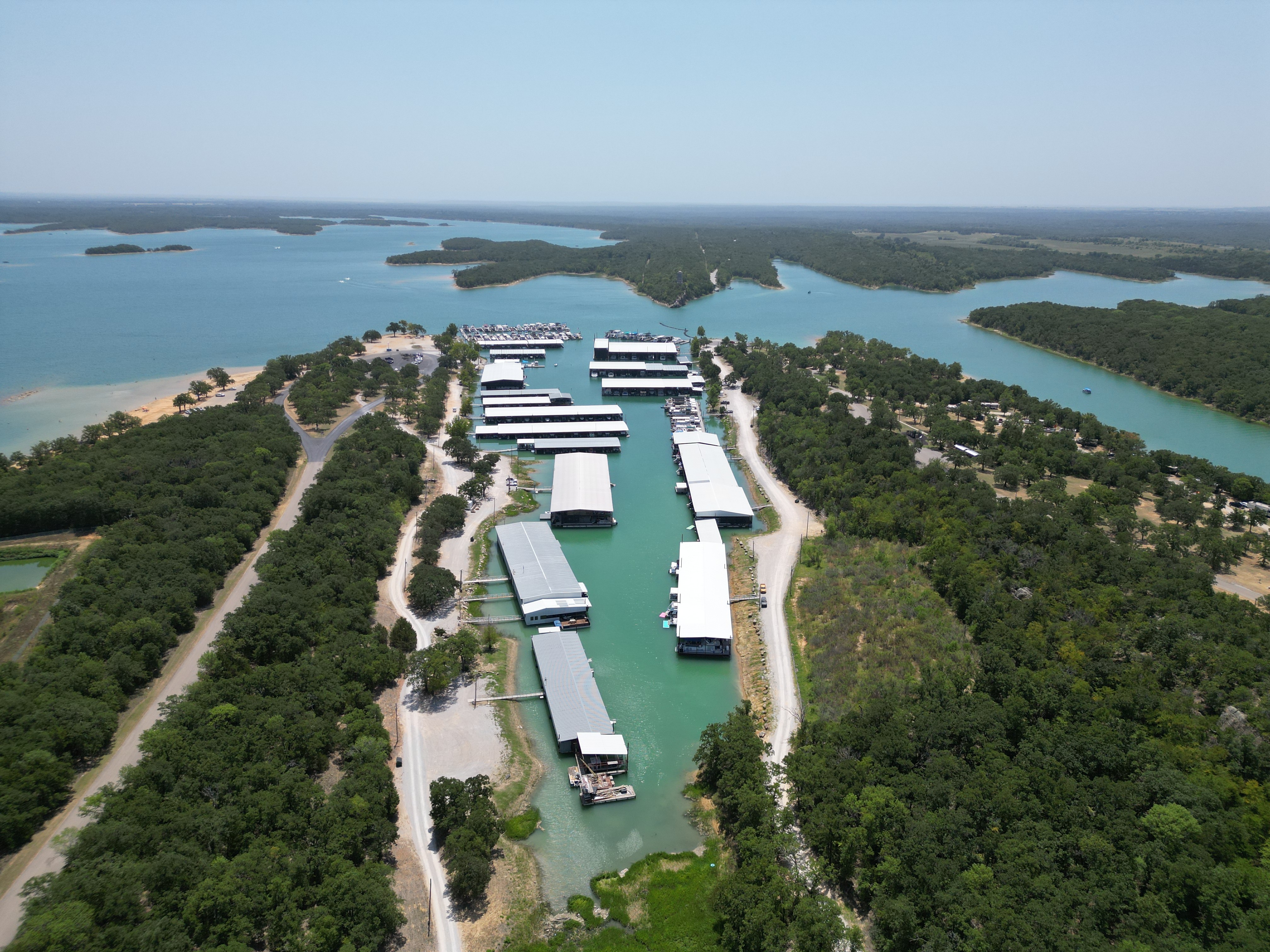 Aerial view of a marina with multiple covered boat docks surrounded by lush greenery, set along a winding body of water under a clear blue sky.