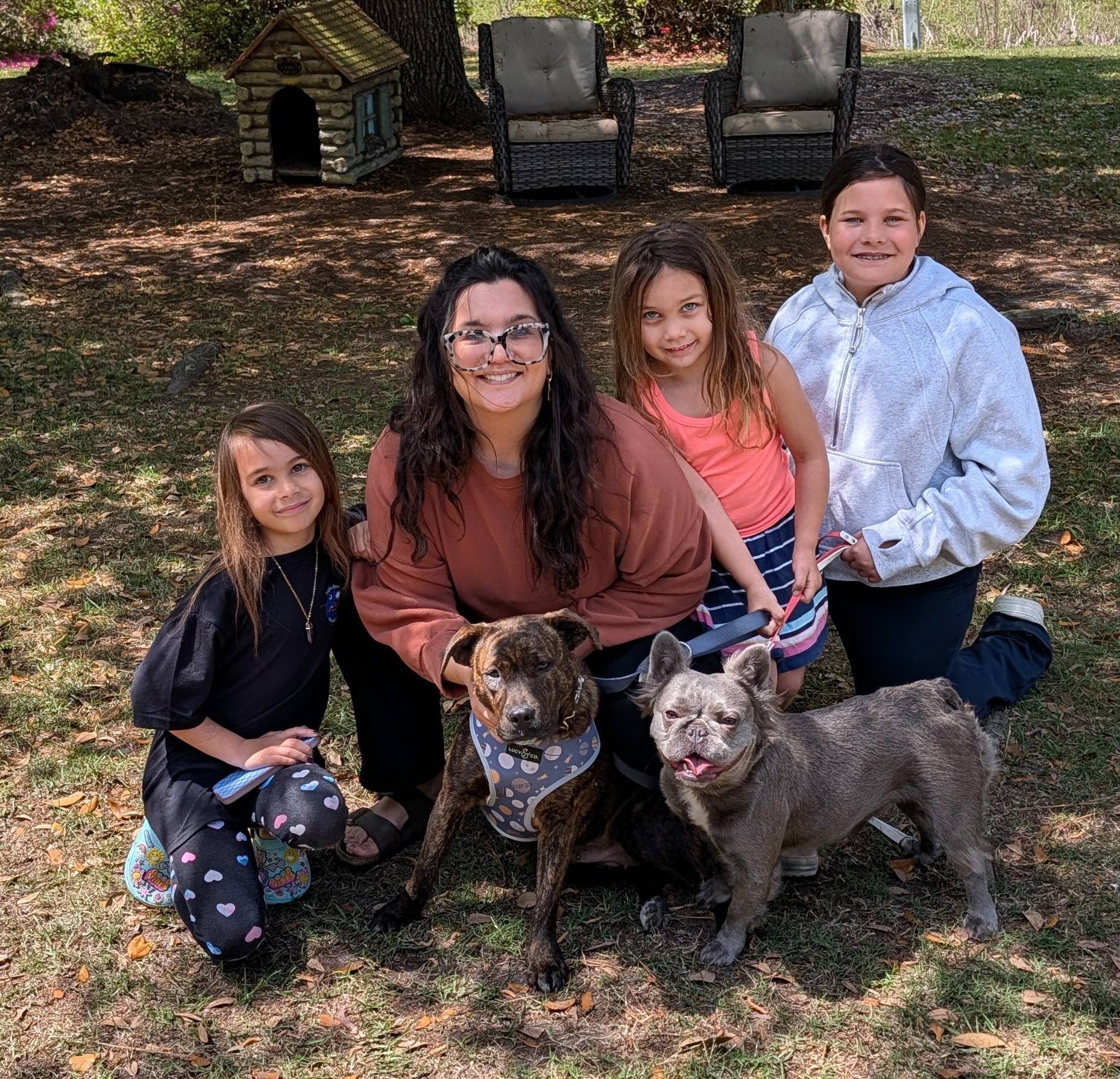 A group of four people holding dogs, smiling outdoors with trees in the background.