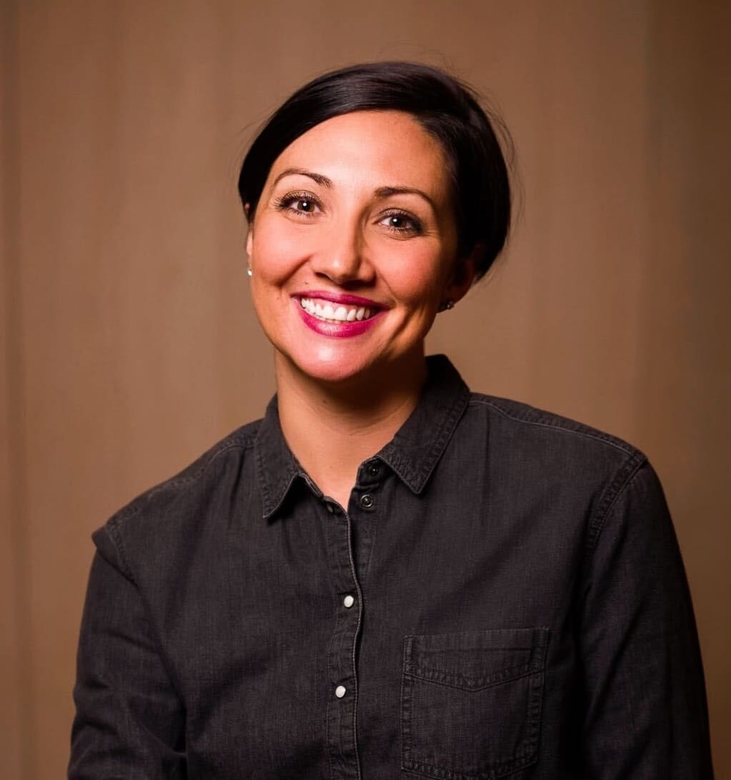 A white woman with short dark hair smiling into camera against brown background