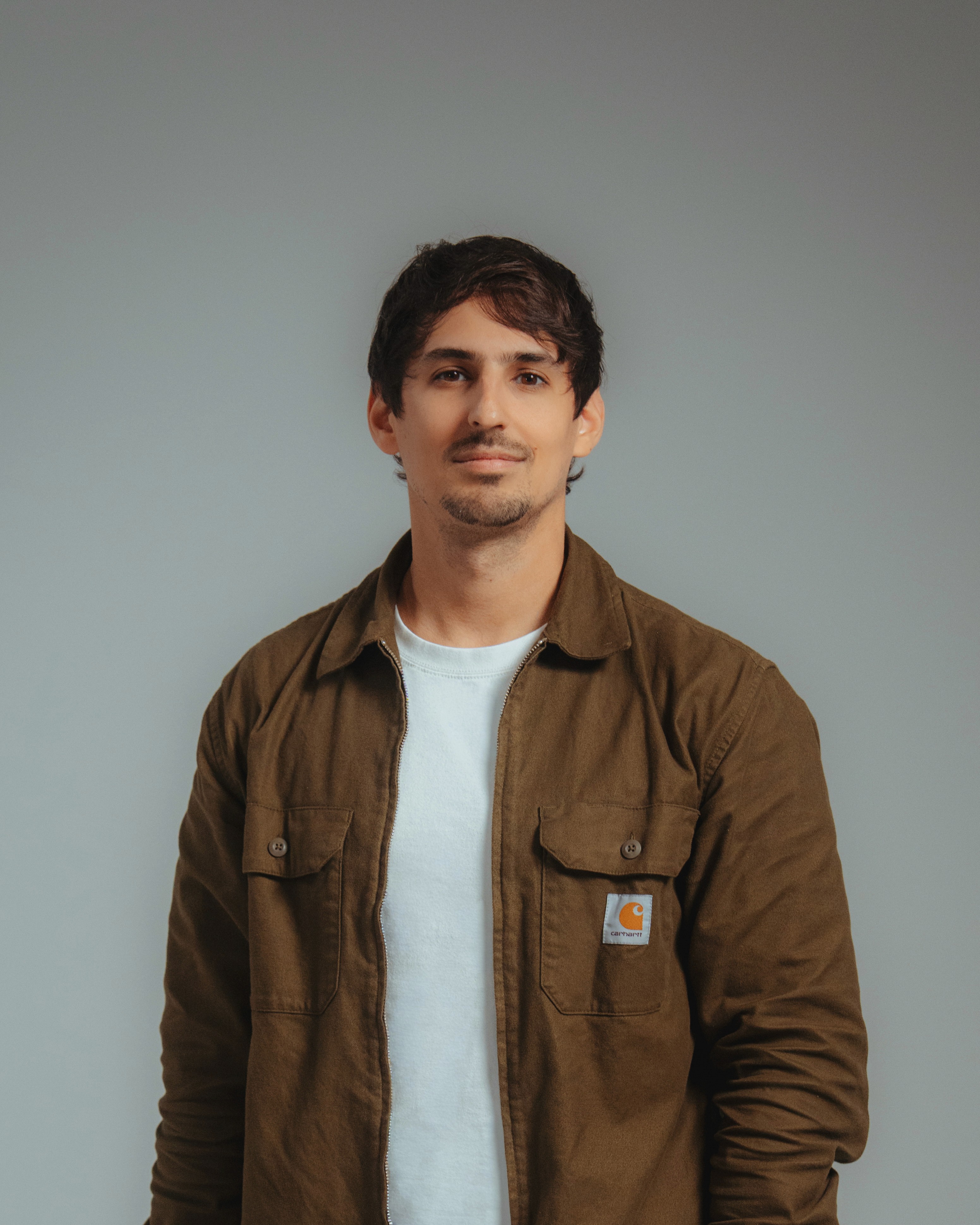 Frontalbild eines jungen Mannes, mit einer braunen Jacke, in die Kamera lächelnd.