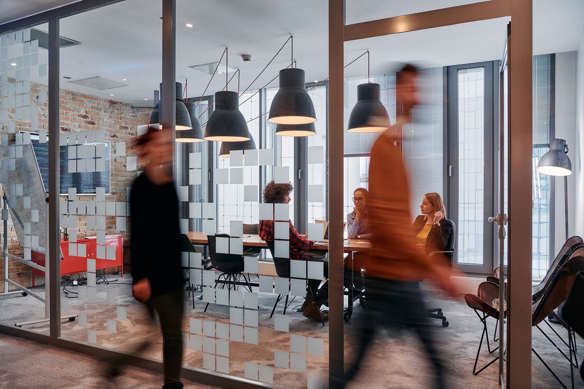 Blurred figures walking in a modern office with meeting room visible, focus on people and work environment.