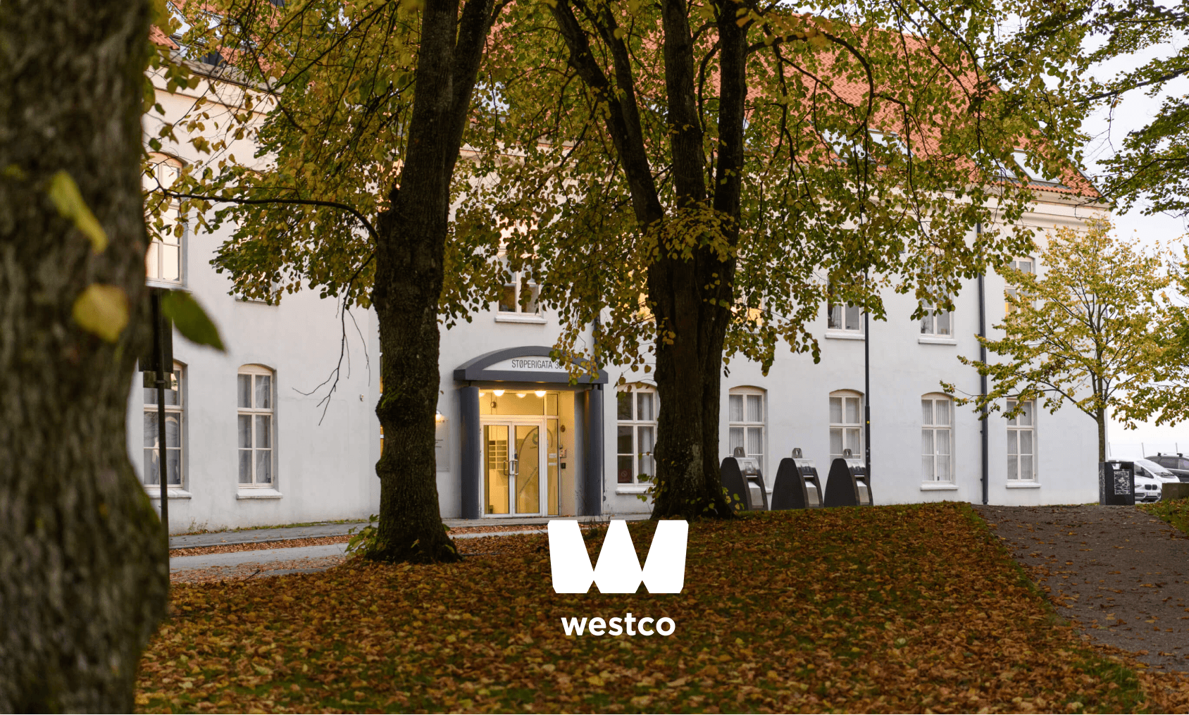 The Westco Eiendom logo overlayed on an image showing the property in Støperigata with trees in front of it. With orange leaves on the ground.