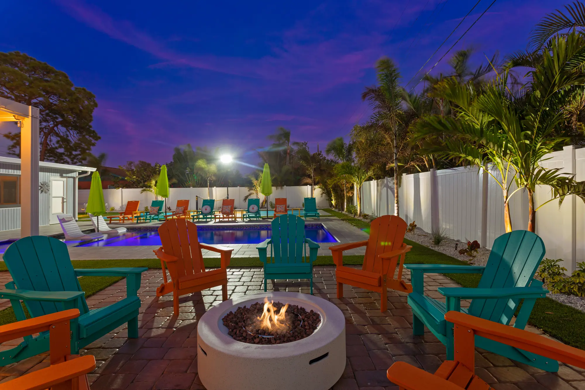 Fire pit ringed by orange and teal Adirondack chairs at twilight, with the pool behind at Pickleball Resort, Bradenton, Florida