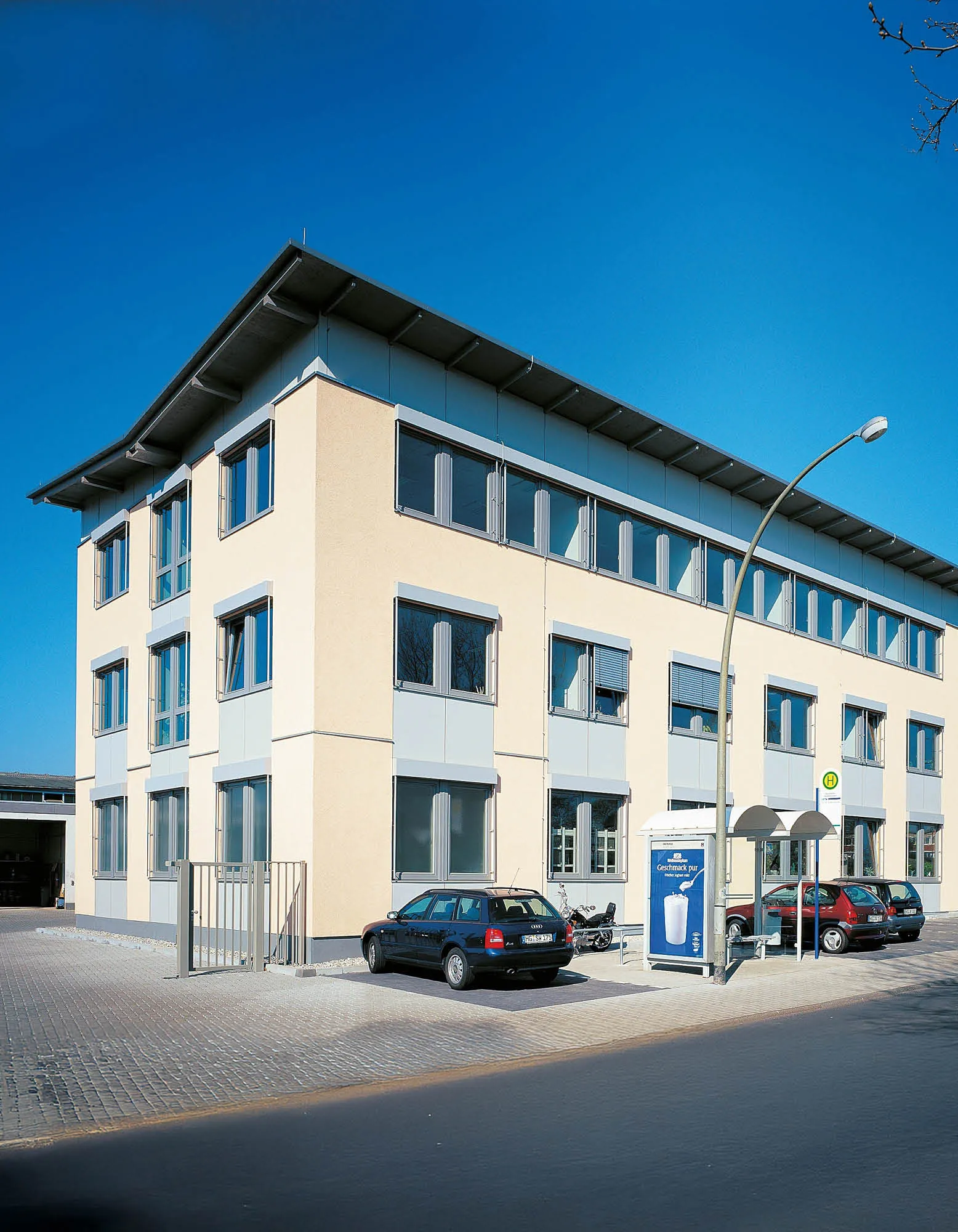 Mehrstöckiges Geschäftsgebäude mit hellbeiger Fassade, vielen Fenstern und einem Parkplatz, an dem mehrere Autos parken.