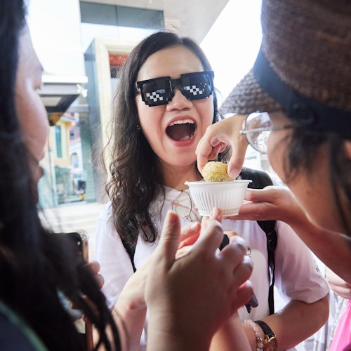 Group of friends sharing food outside; central person wearing pixelated sunglasses excitedly opening mouth near white bowl.