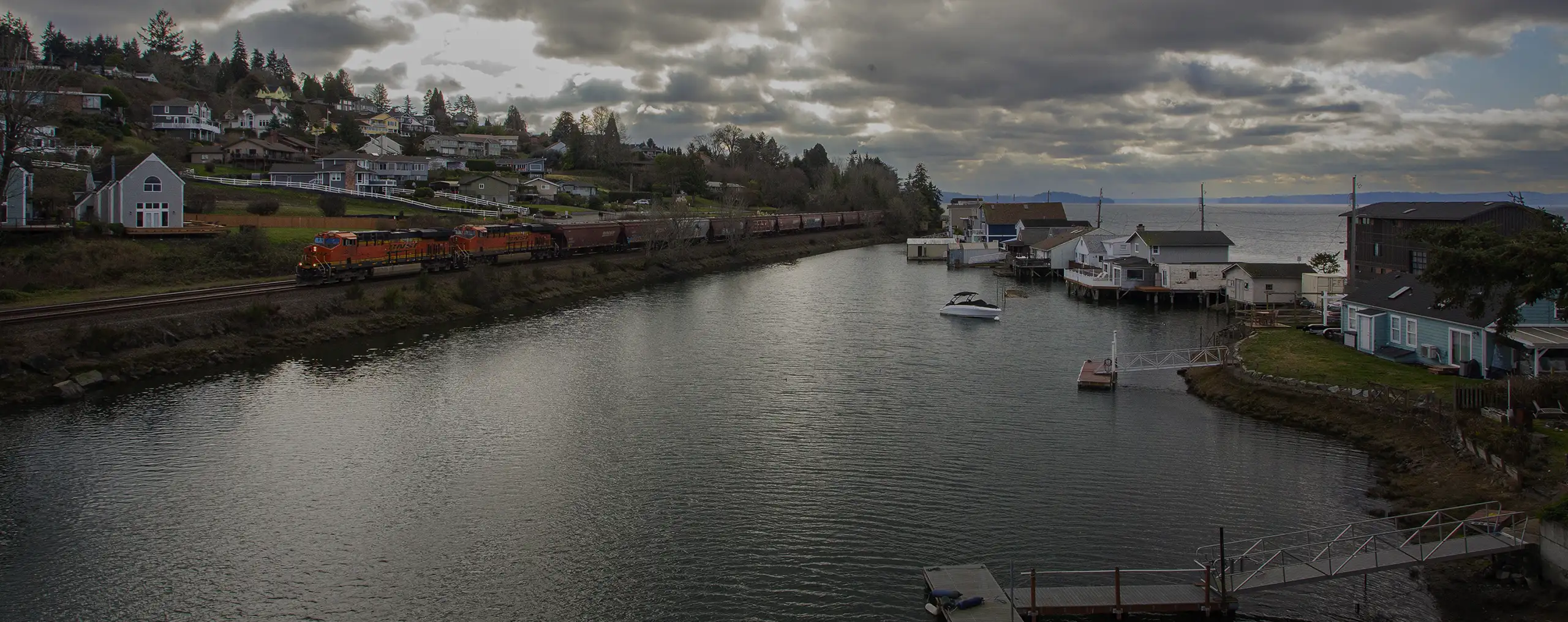 An image of the City of University Place along the water. There are floating houses to the right, the shore is on the left, and a freight train is going along the edge and divides the land and water. Residential houses populate the land and goes up a hill with trees, and there are a few boats and docks extending from the floating houses on the water.