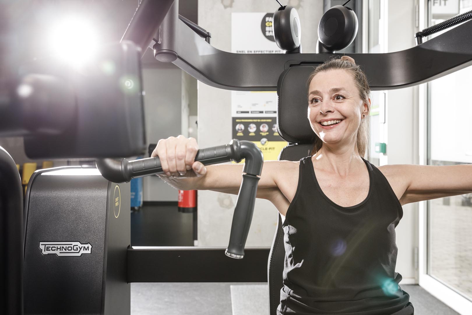 A older woman smiling while exercising on a gym machine, with bright lights in the background.