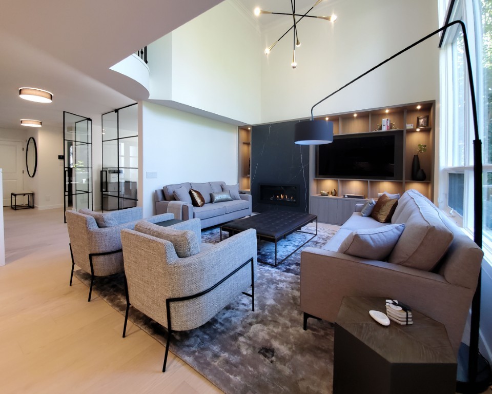A living room with warm light flooring, a grey rug, and semi matching furniture. Illuminated custom millwork and a grand fireplace are in the background. An open glass door is to the left.
