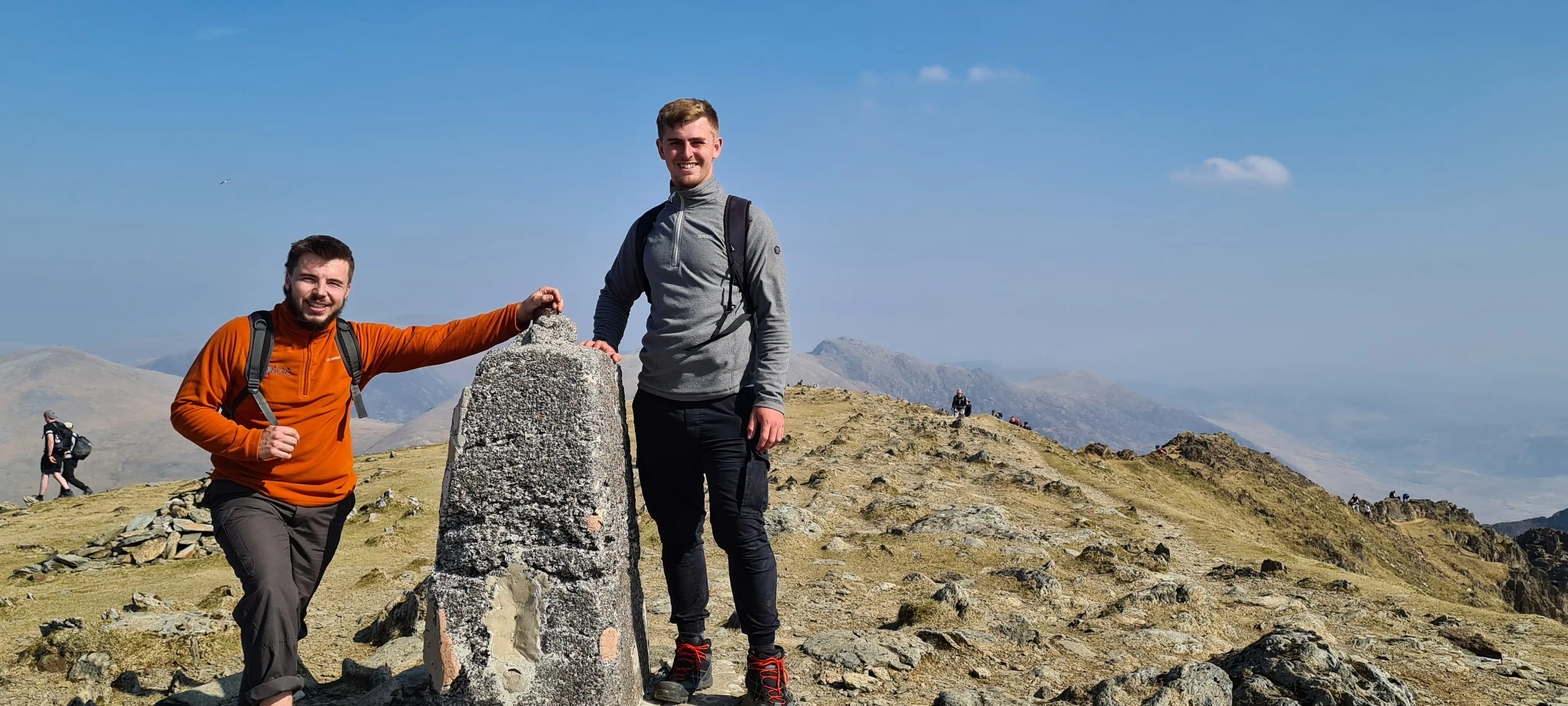 Two hikers stand on a mountain peak, one in an orange jacket and the other in a gray shirt, with a clear blue sky.
