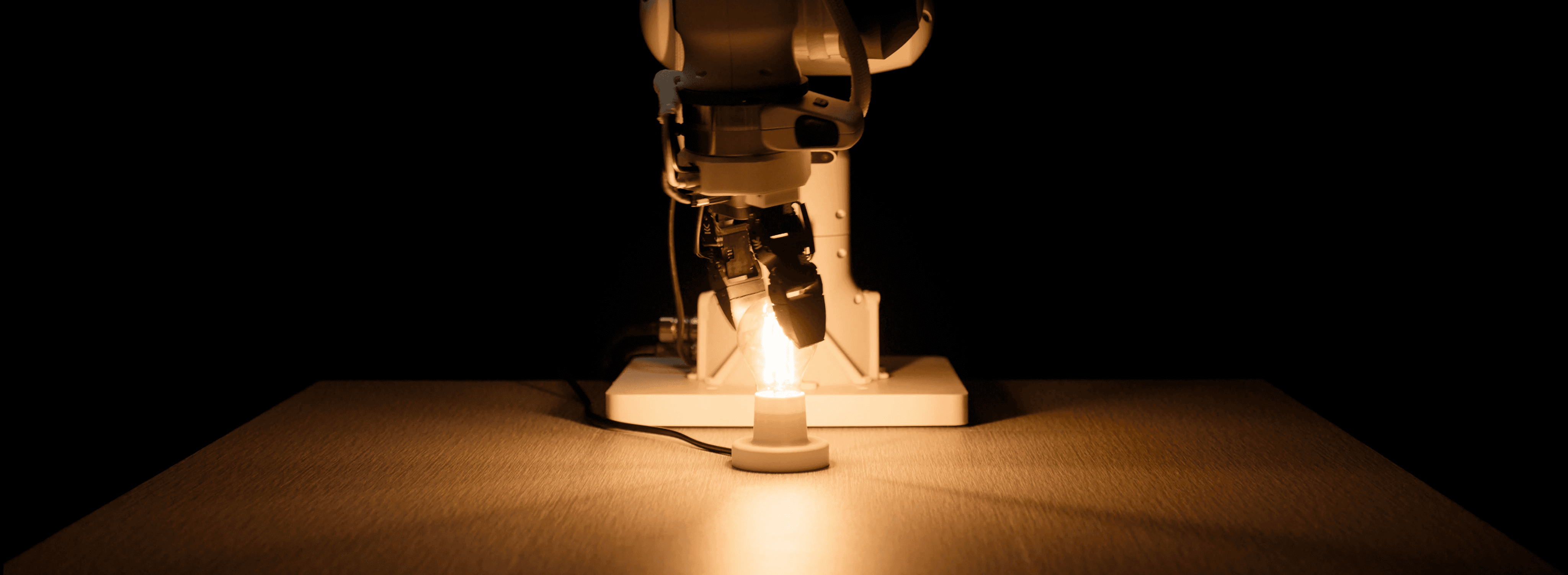 An Eka Robotics arm and gripper screwing a glowing light bulb into a socket on a table in a dark room.