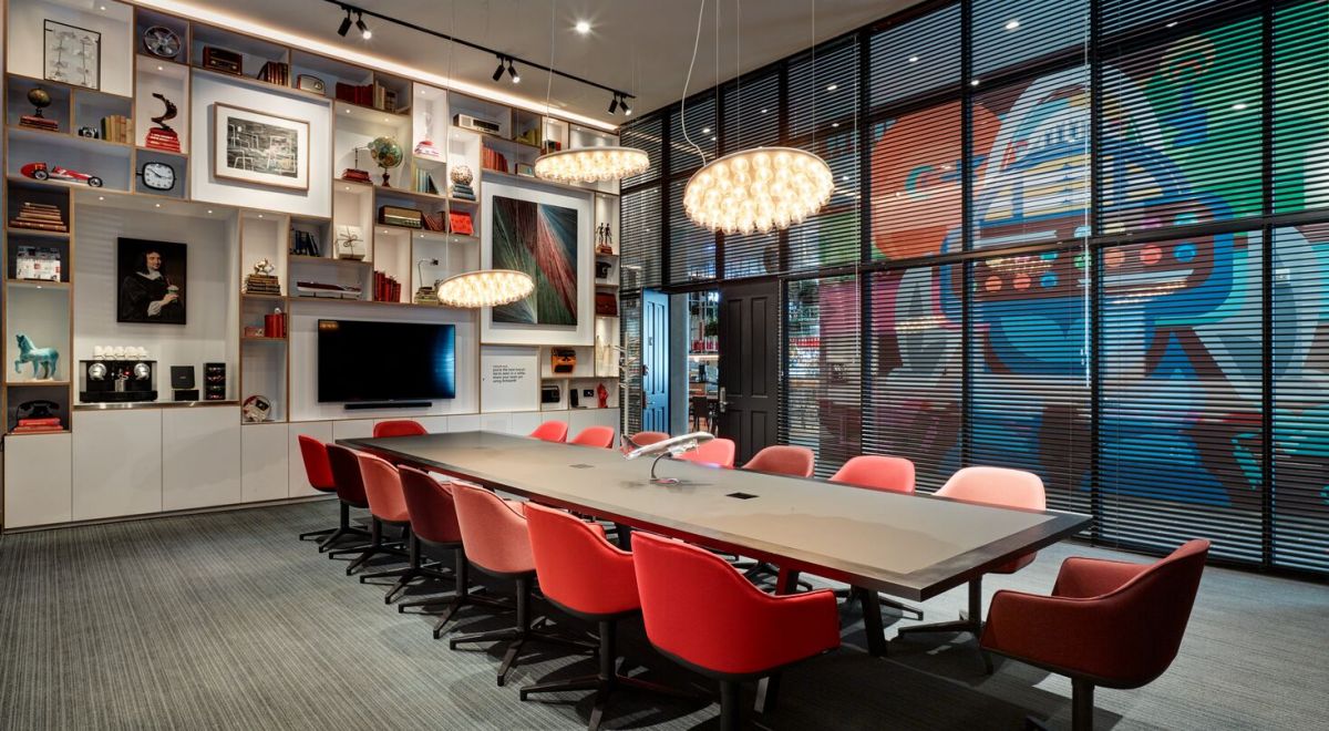 Ground-floor conference room with floor-to-ceiling glazing, a spacious table surrounded by red chairs, and an eclectic built-in bookcase showcasing quirky art and artifacts.