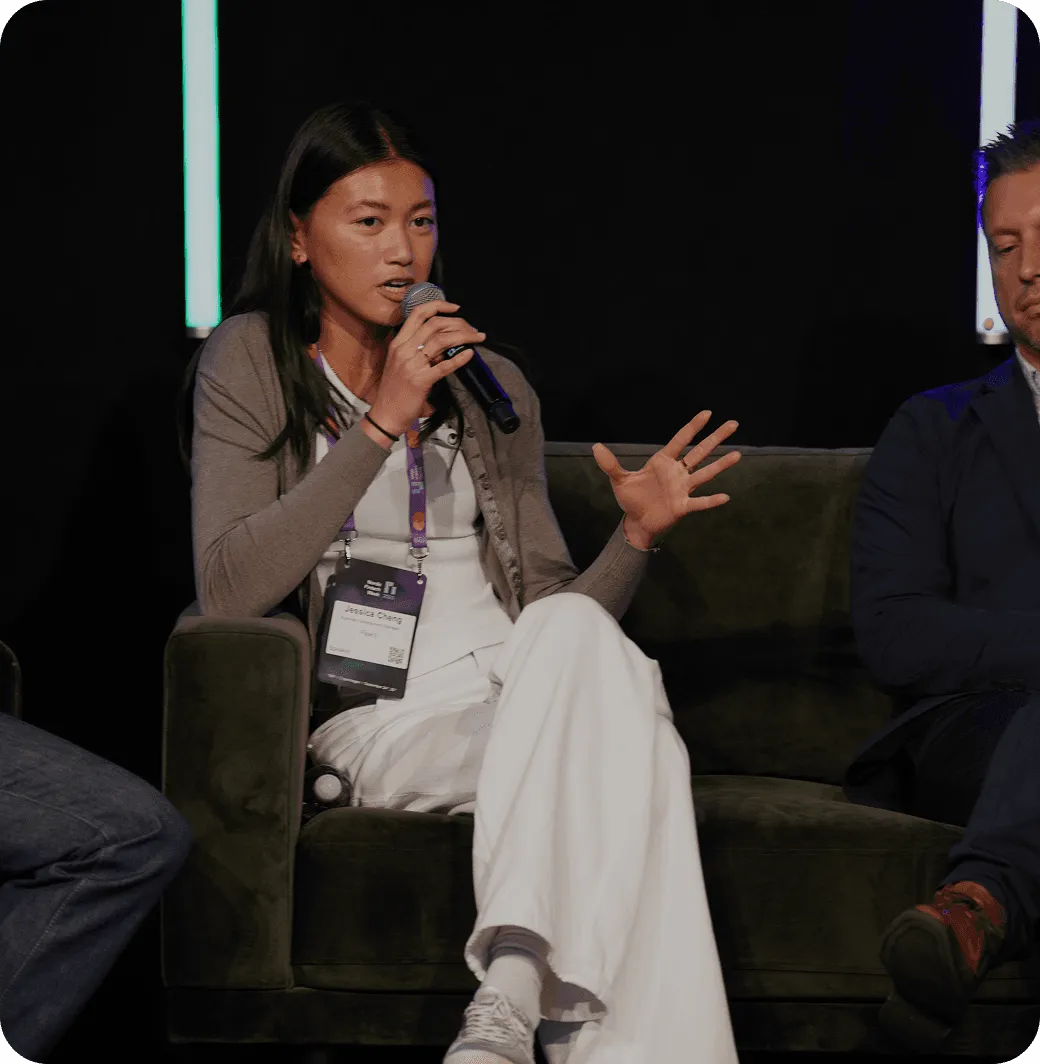 A woman speaks into a microphone while seated on a couch, flanked by two men during a panel discussion.