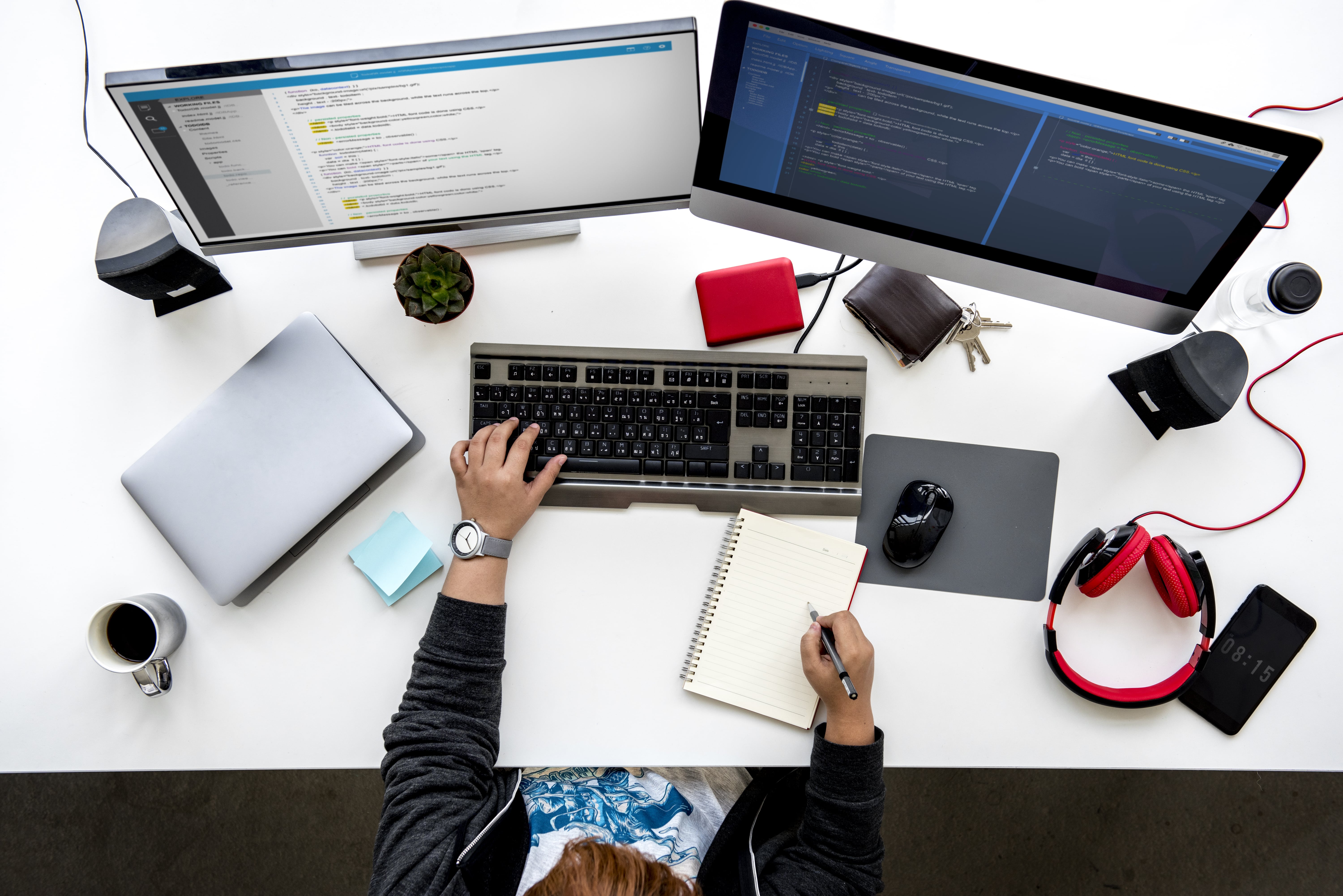 Top-down view of a web developer's desk with a dual-monitor setup showing code, a person typing on a keyboard, and writing in a notebook.