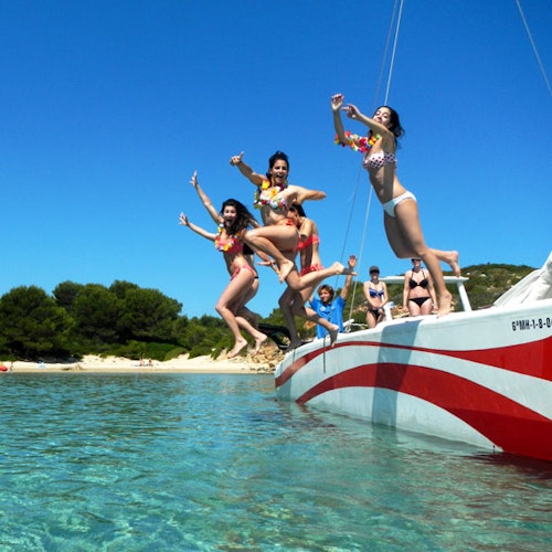 Ljudi u kupaćim kostimima skaču s broda s crvenim prugama u čisto plavo more blizu plaže s drvećem pod vedrim nebom.