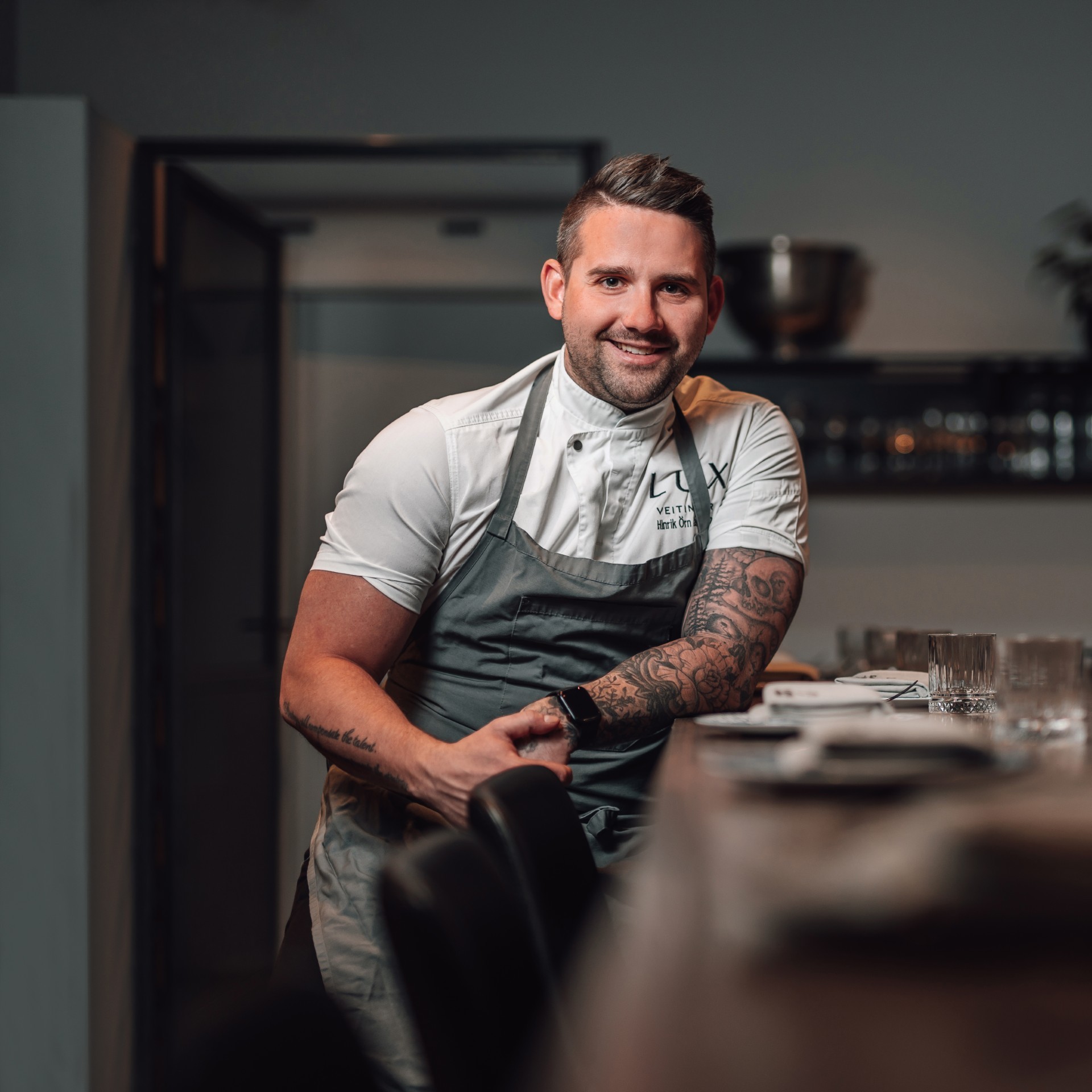Icelandic chef Hinrik Örn Lárusson leans against the end of a set dining table, smiling at the camera in a white chef's jacket and grey Lux Veitingar apron, with a tattooed forearm visible.