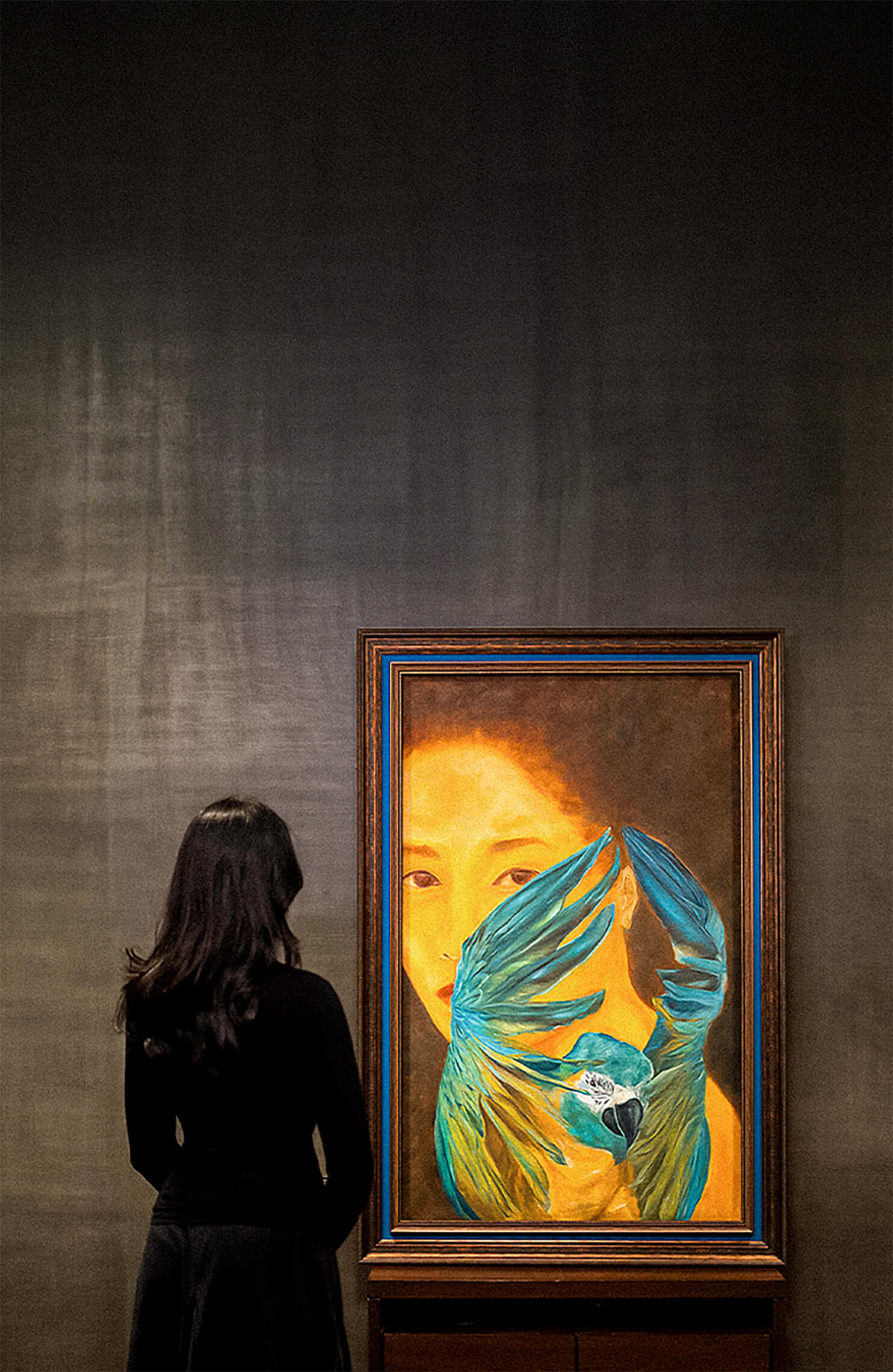 Person viewing a framed painting of a woman’s face with blue bird wings and a parrot in an art gallery.