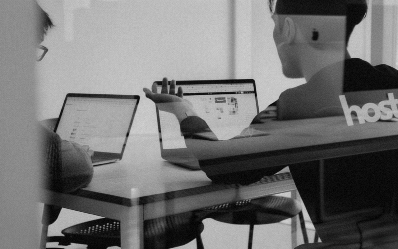 Black and white image of people working at a table with laptops, showing hands gesturing during discussion of digital content displayed on screens, with fabrica® logo in the corner.